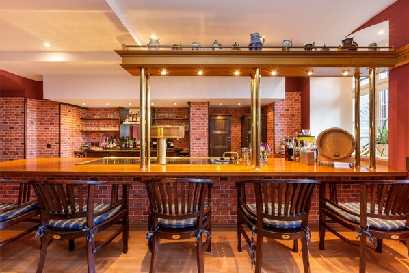 A bar area with a long wooden counter, four wooden bar stools with blue cushions, and a brick wall backdrop. - Schlosshotel Ralswiek (Ralswiek)