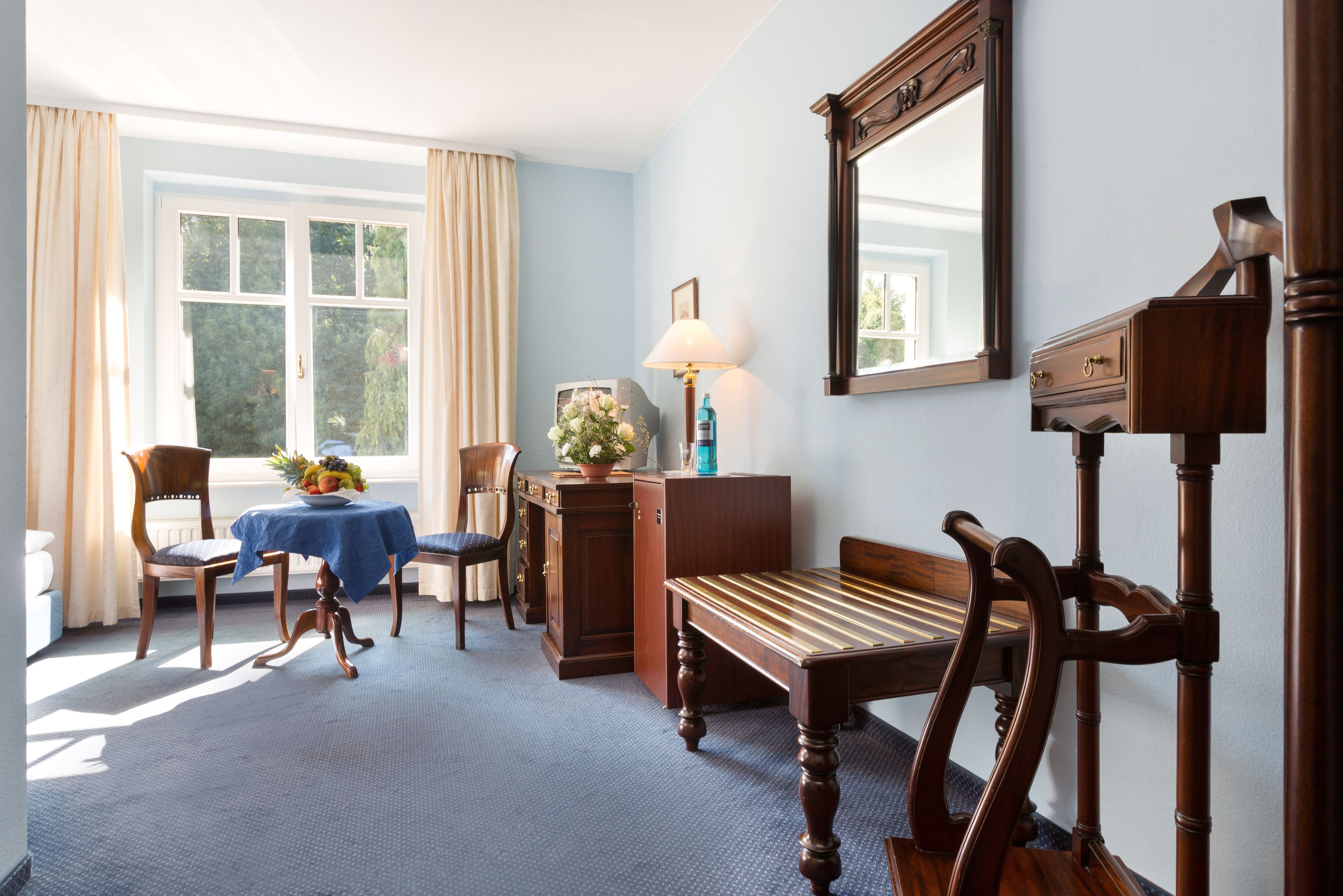 A room with a wooden bench, a table with a blue tablecloth, a lamp, a mirror, and a window with curtains.