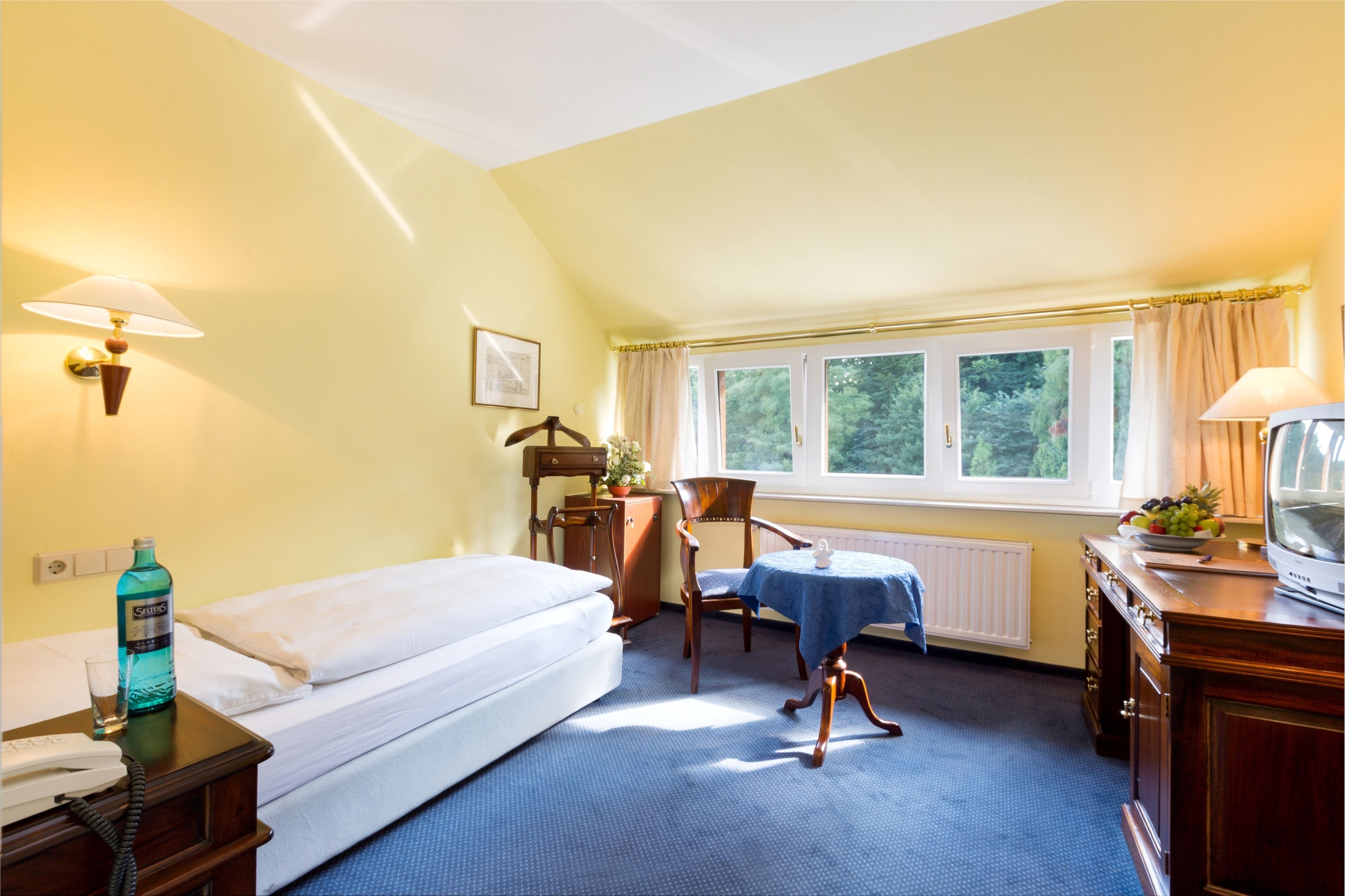A hotel room with a bed, a desk with a telephone, a chair, a table with a tablecloth, a radiator, and a window with curtains.