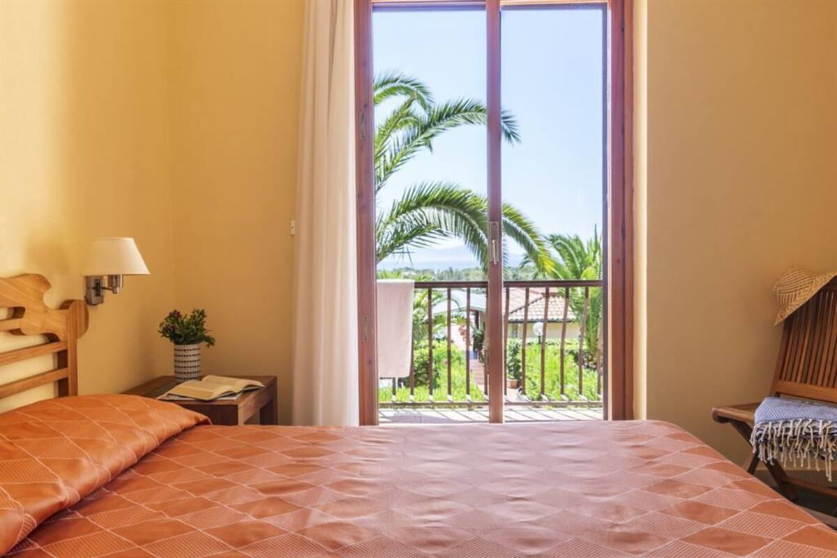 Un dormitorio con cama, mesita de noche, lámpara, ventana con vista a palmeras y balcón.