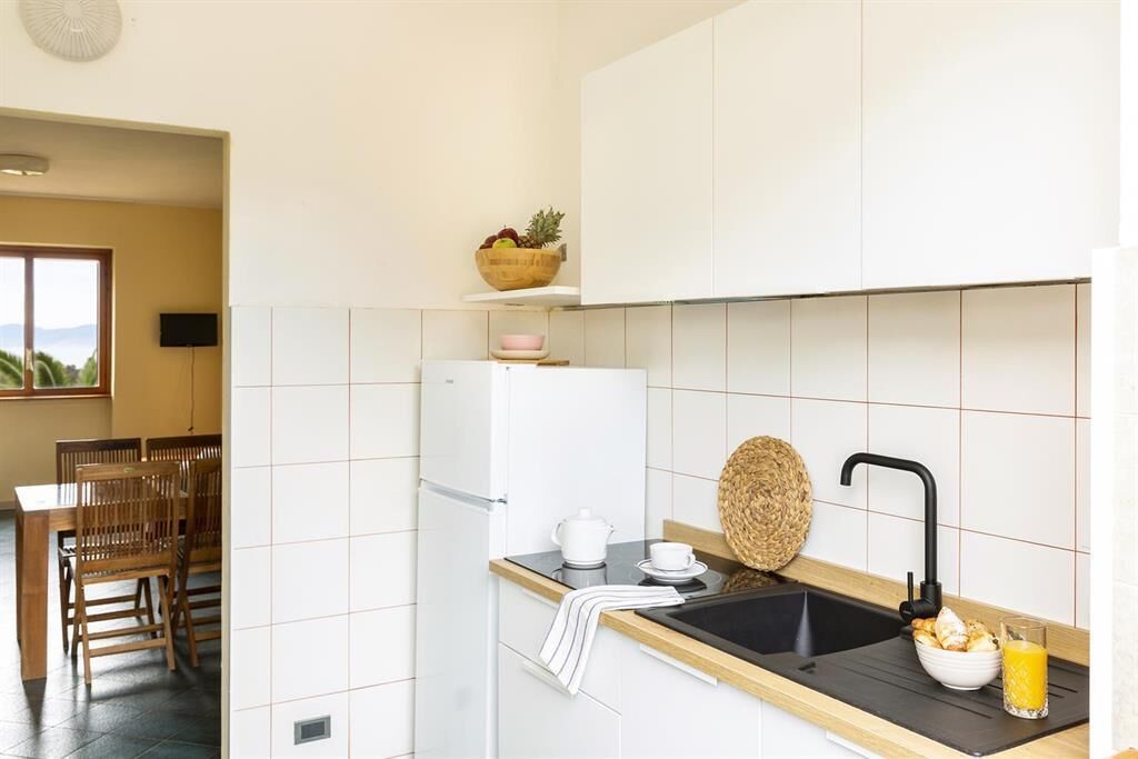 Eine Küche mit weißen Schränken, einer schwarzen Arbeitsplatte, einem Waschbecken mit schwarzem Wasserhahn, einer Obstschale auf der Arbeitsplatte sowie ein Essbereich mit Holztischen und einer Tischplatte.