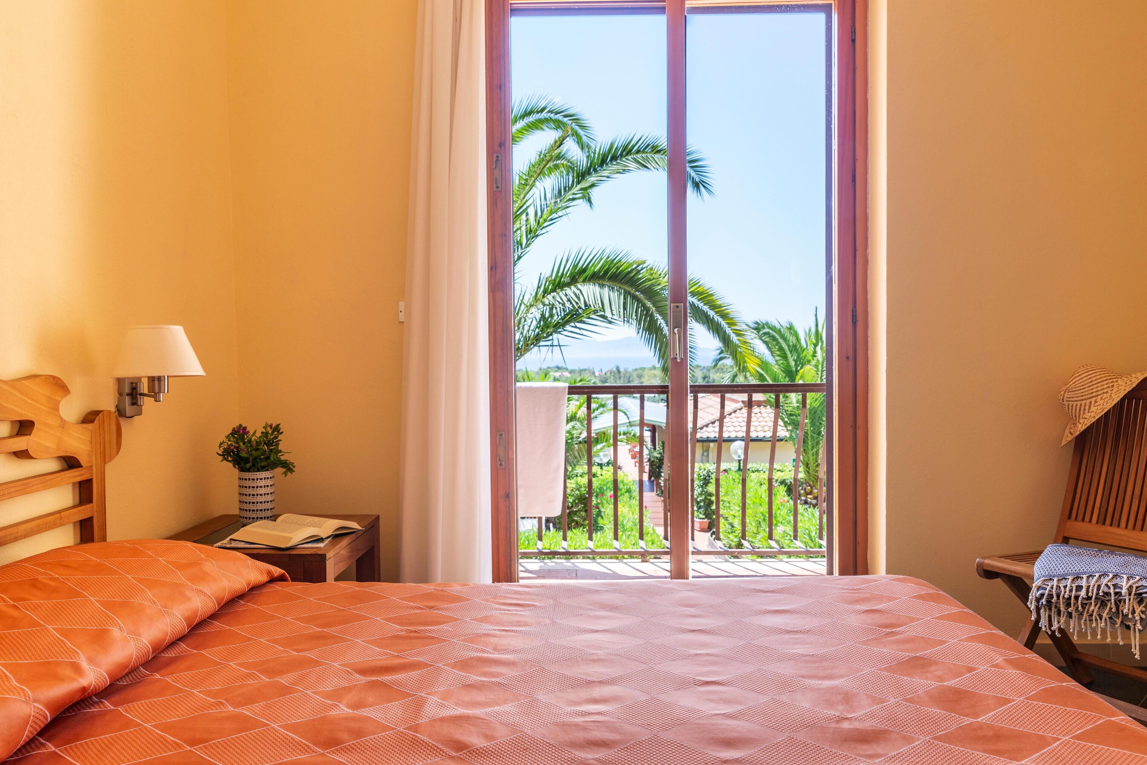 Une chambre à coucher avec un lit, une table de chevet, une lampe et une vue sur un balcon agrémenté de palmiers.