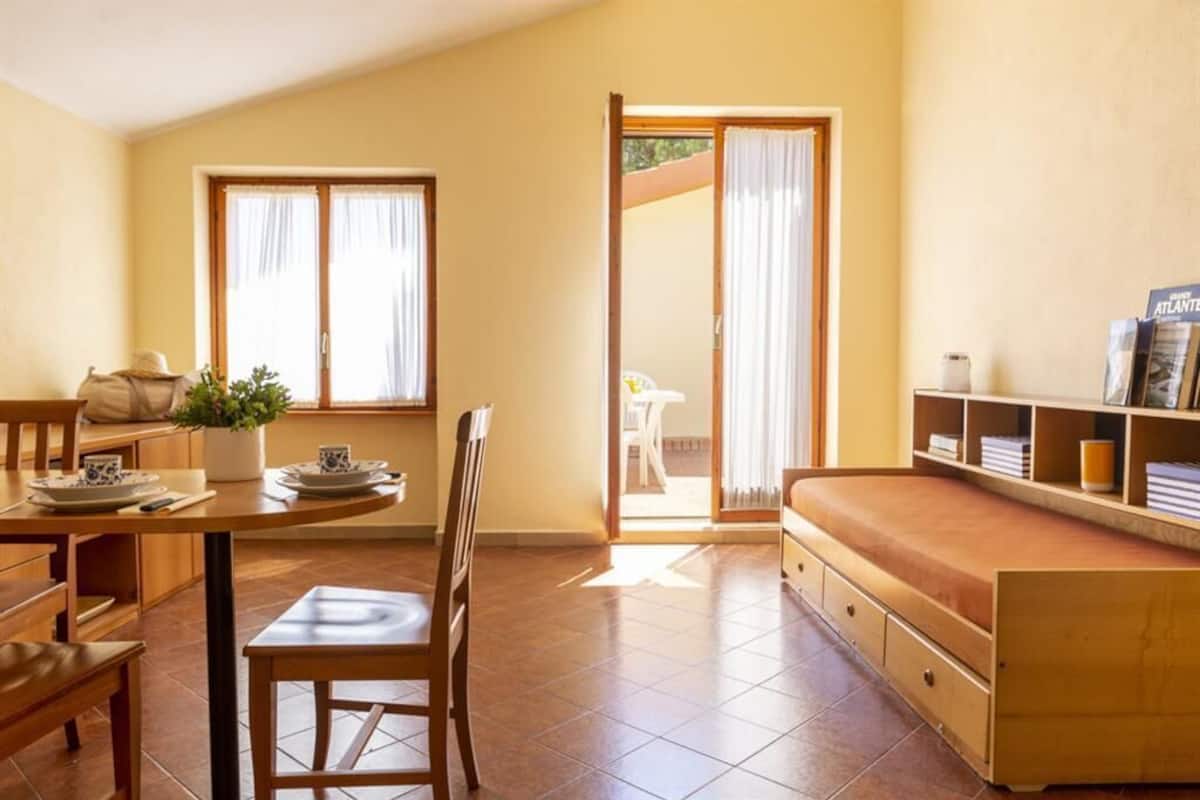 Una habitación acogedora con una cama de madera, una mesa de comedor con sillas, una pequeña cocina y un ventanal corredizo que da a un espacio exterior.