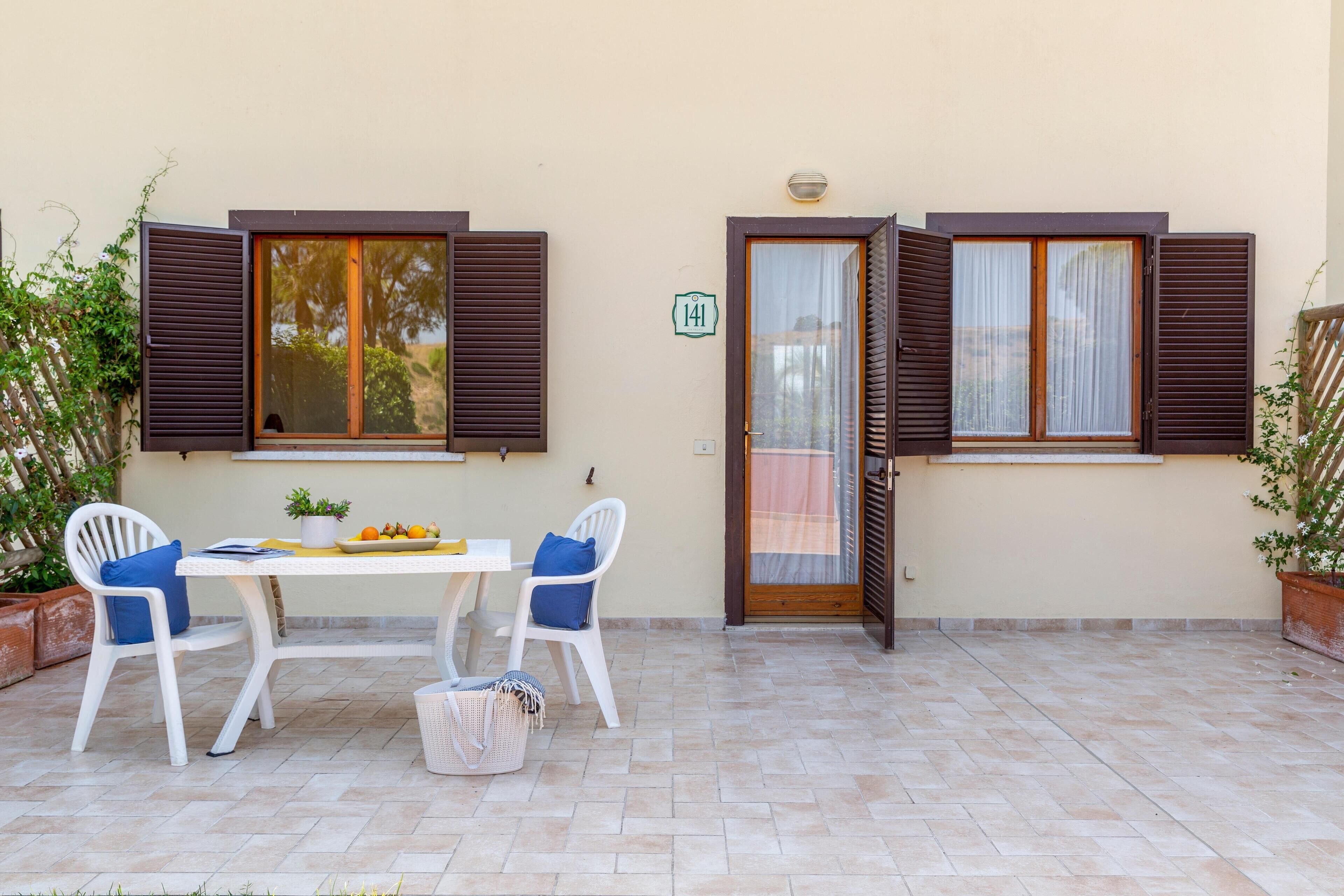 A patio with a table and chairs, a door with a number sign, and a window with shutters.