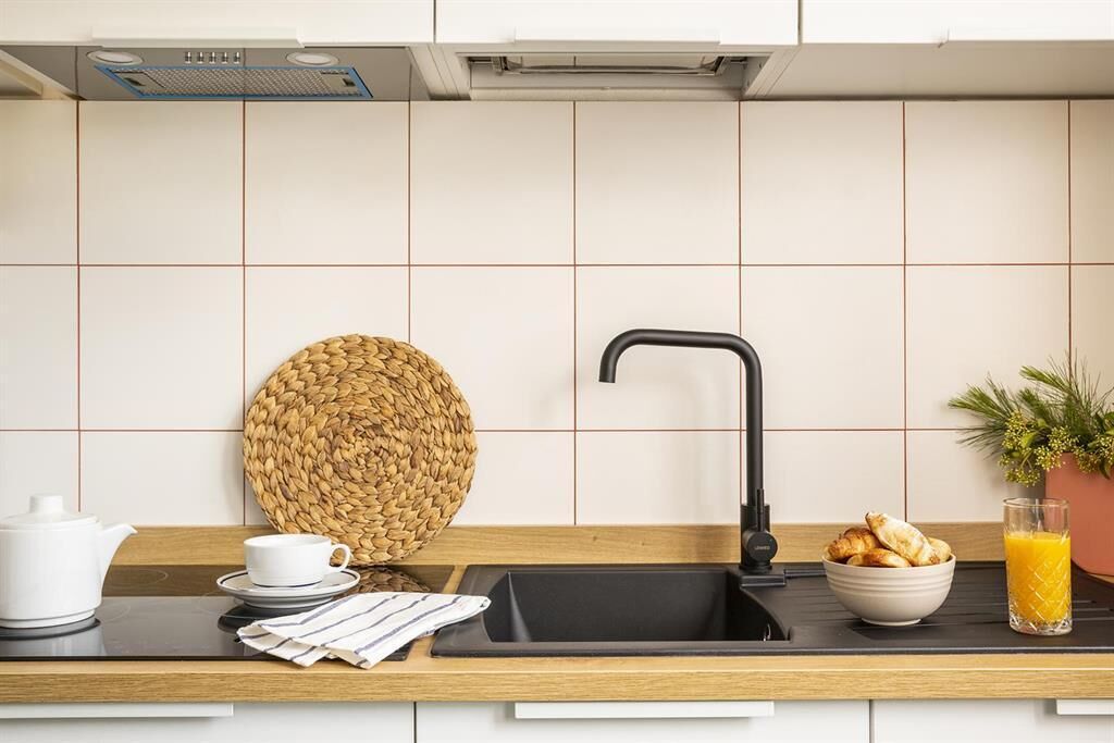 Une cuisine avec un évier noir, un panier en osier, une théière blanche, une tasse, un bol de croissants et un verre de jus d’orange.