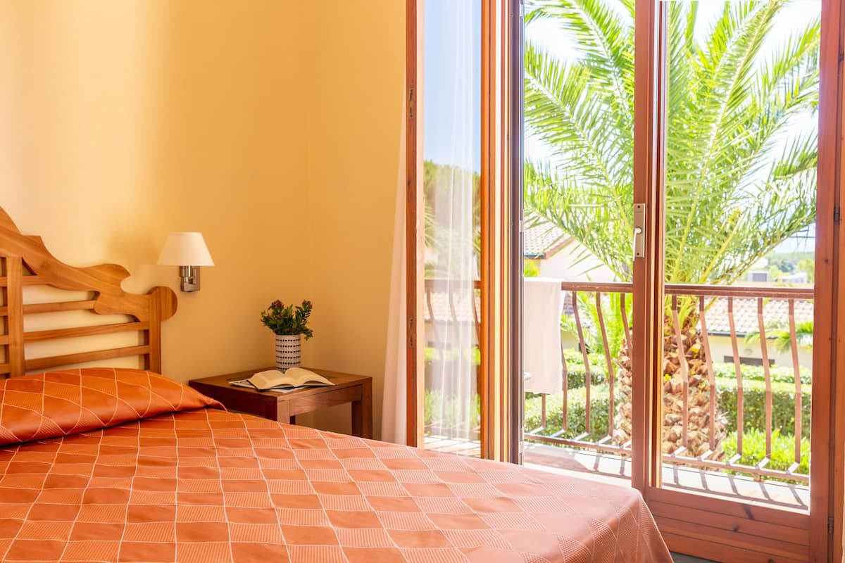 Un dormitorio con cama, mesita de noche, lámpara y vistas a un balcón con palmeras.