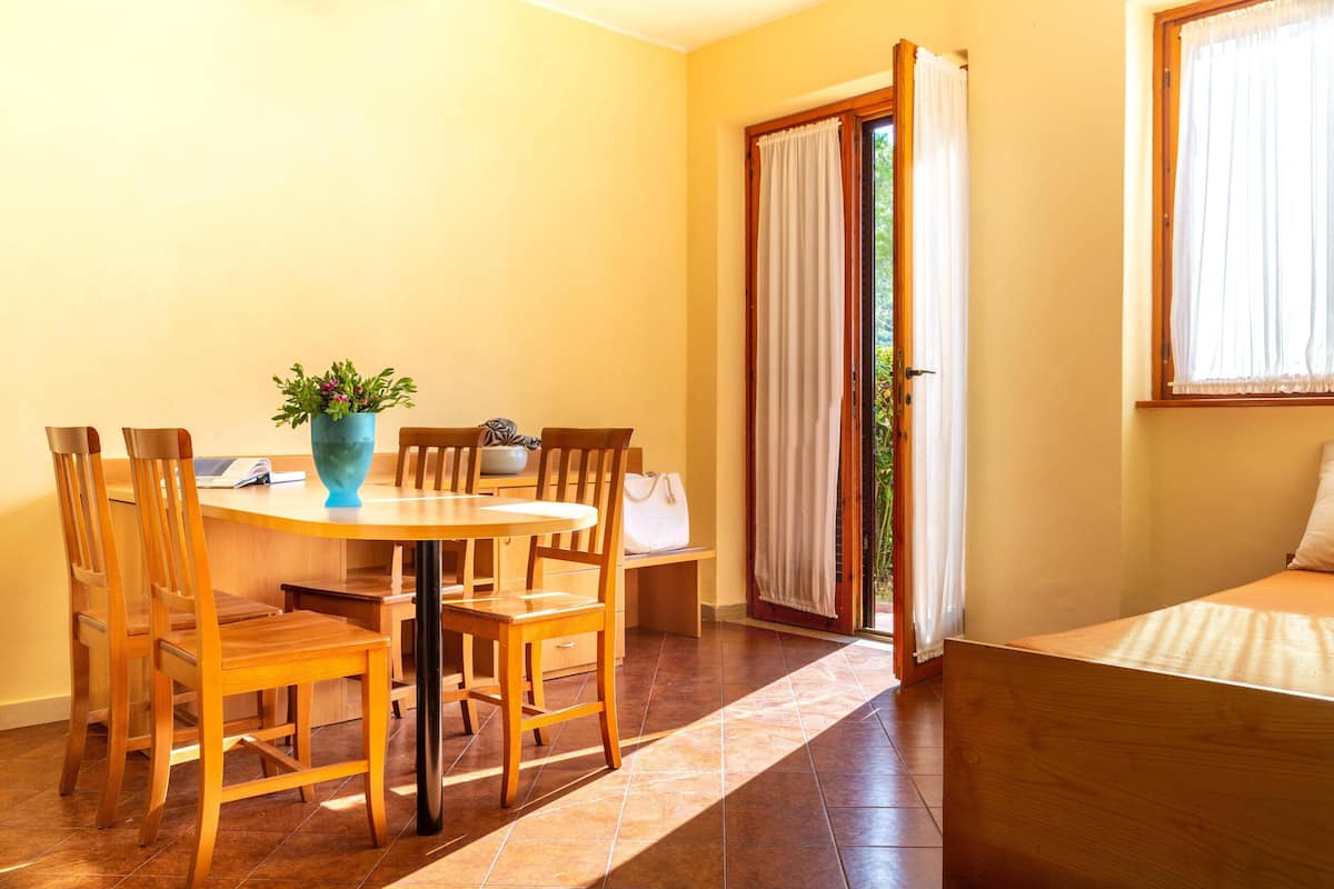 Una habitación con una mesa de comedor de madera, sillas, un jarrón azul con flores, una cama con cubrecama blanco y un ventanal corredizo que da a un espacio exterior.