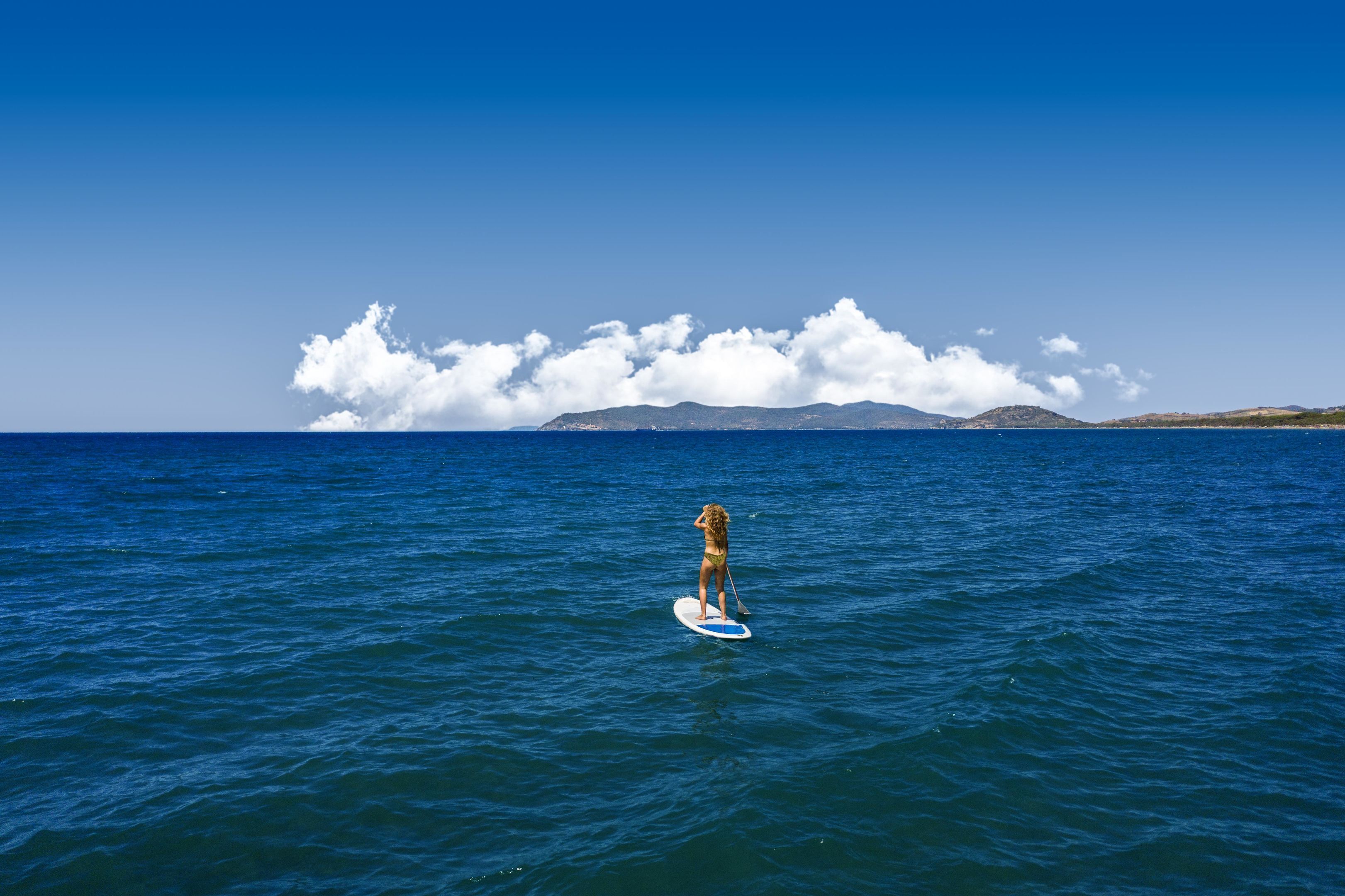 Une personne fait du paddle sur une mer calme, sous un ciel dégagé, avec une terre visible au loin.