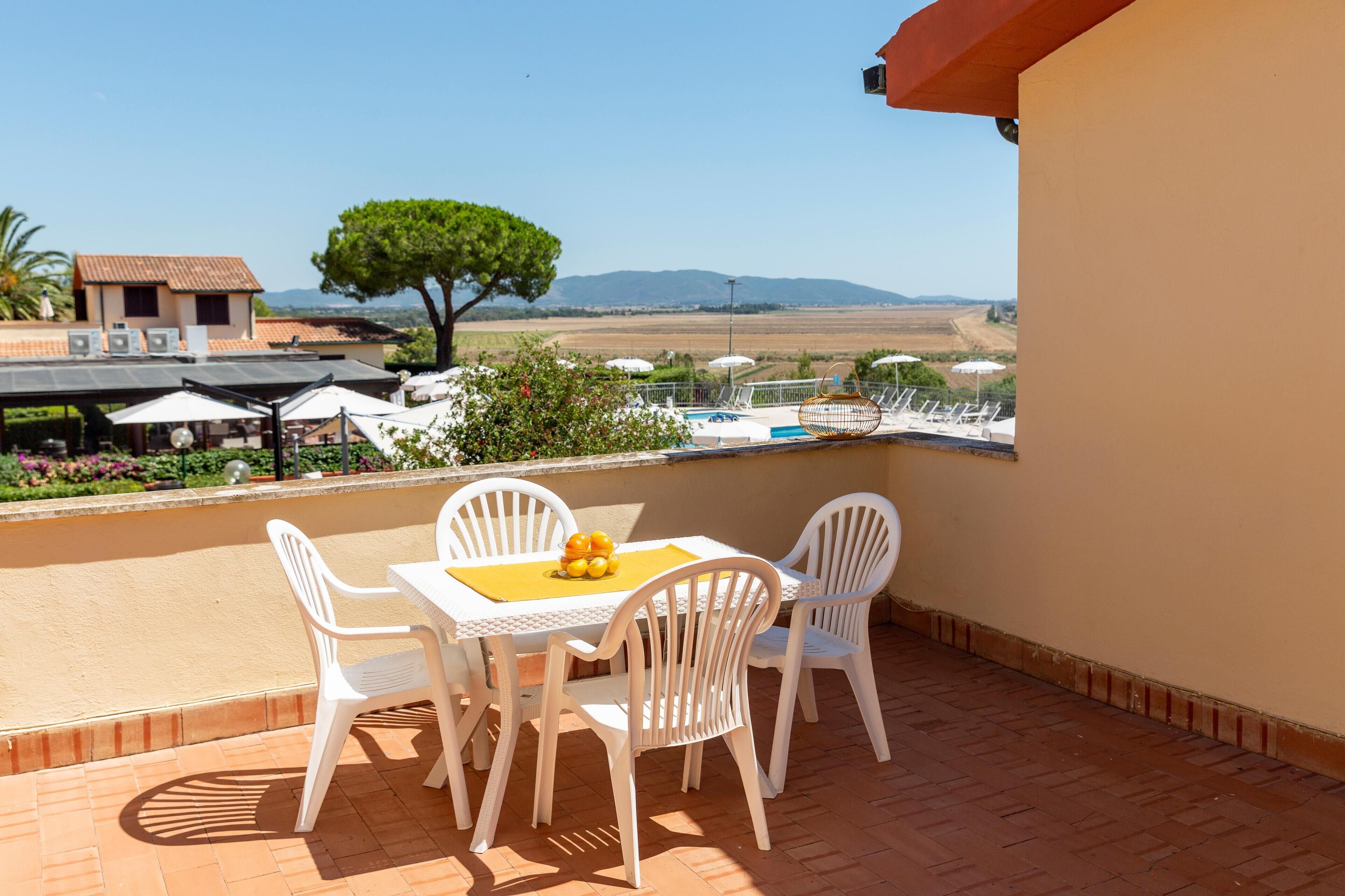 Une terrasse avec des chaises et une table en plastique blanc, donnant sur une piscine et un paysage pittoresque.