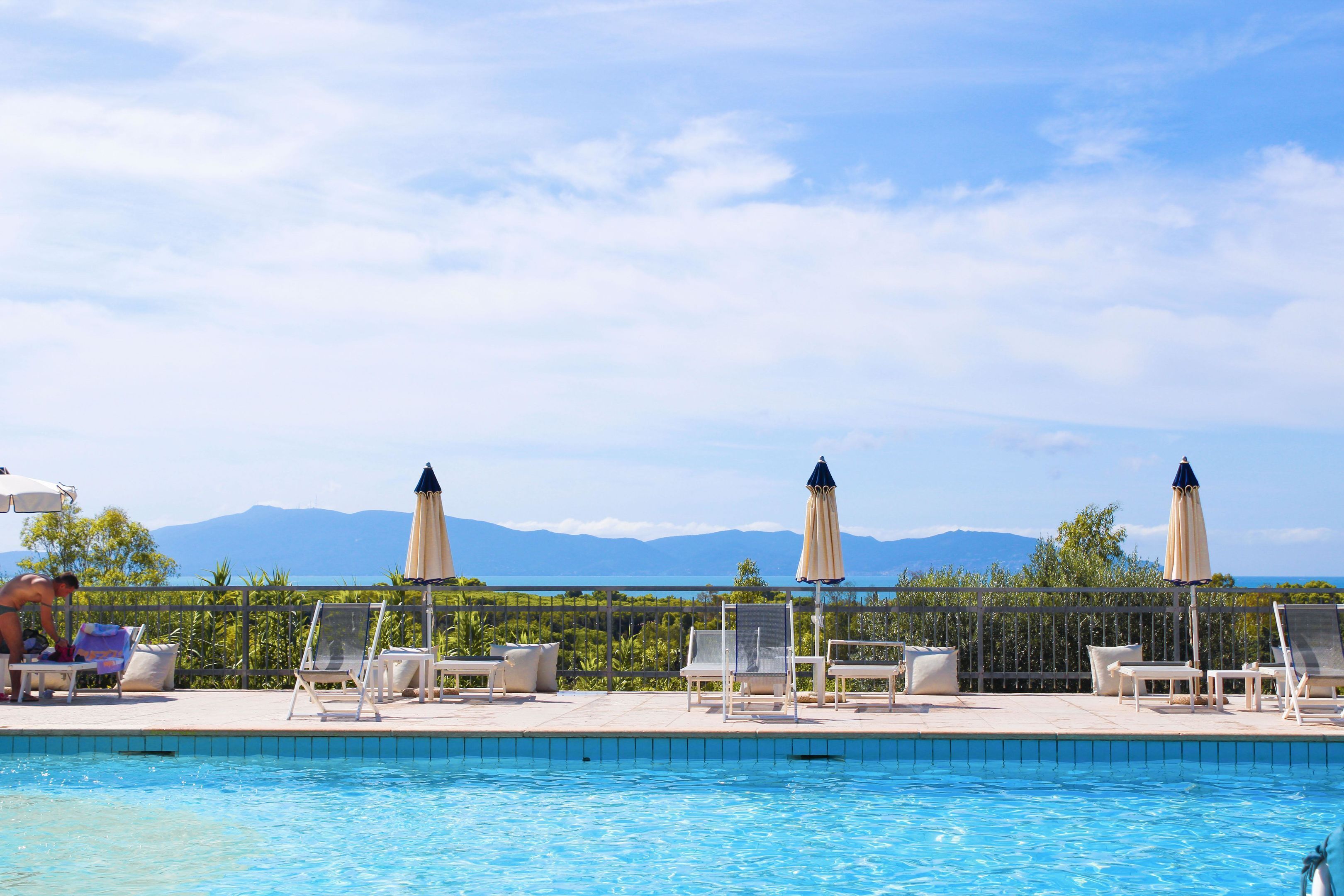 Zona de piscina con sillones de descanso y sombrillas, cielo despejado y montañas al fondo.