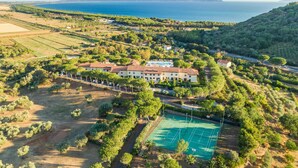 Luchtbeeld van een resort met een tennisbaan, weelderige begroeiing en uitzicht op zee.