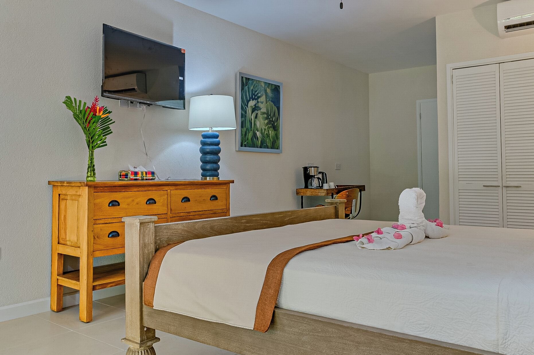 A bedroom with a bed, a wooden dresser, a TV mounted on the wall, a painting, and a white door.