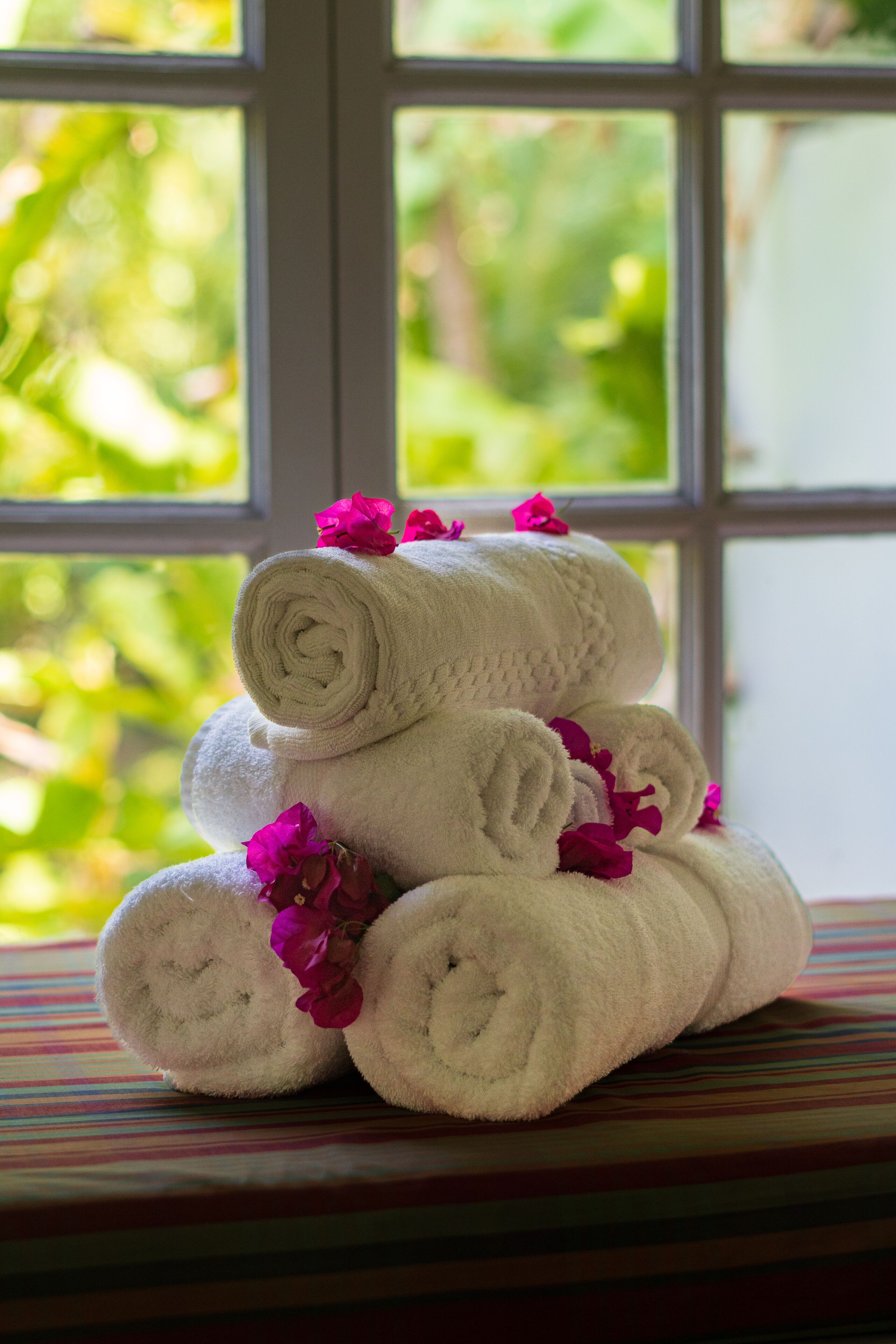 A stack of white towels with pink flowers on a striped surface by a window with a view of greenery.