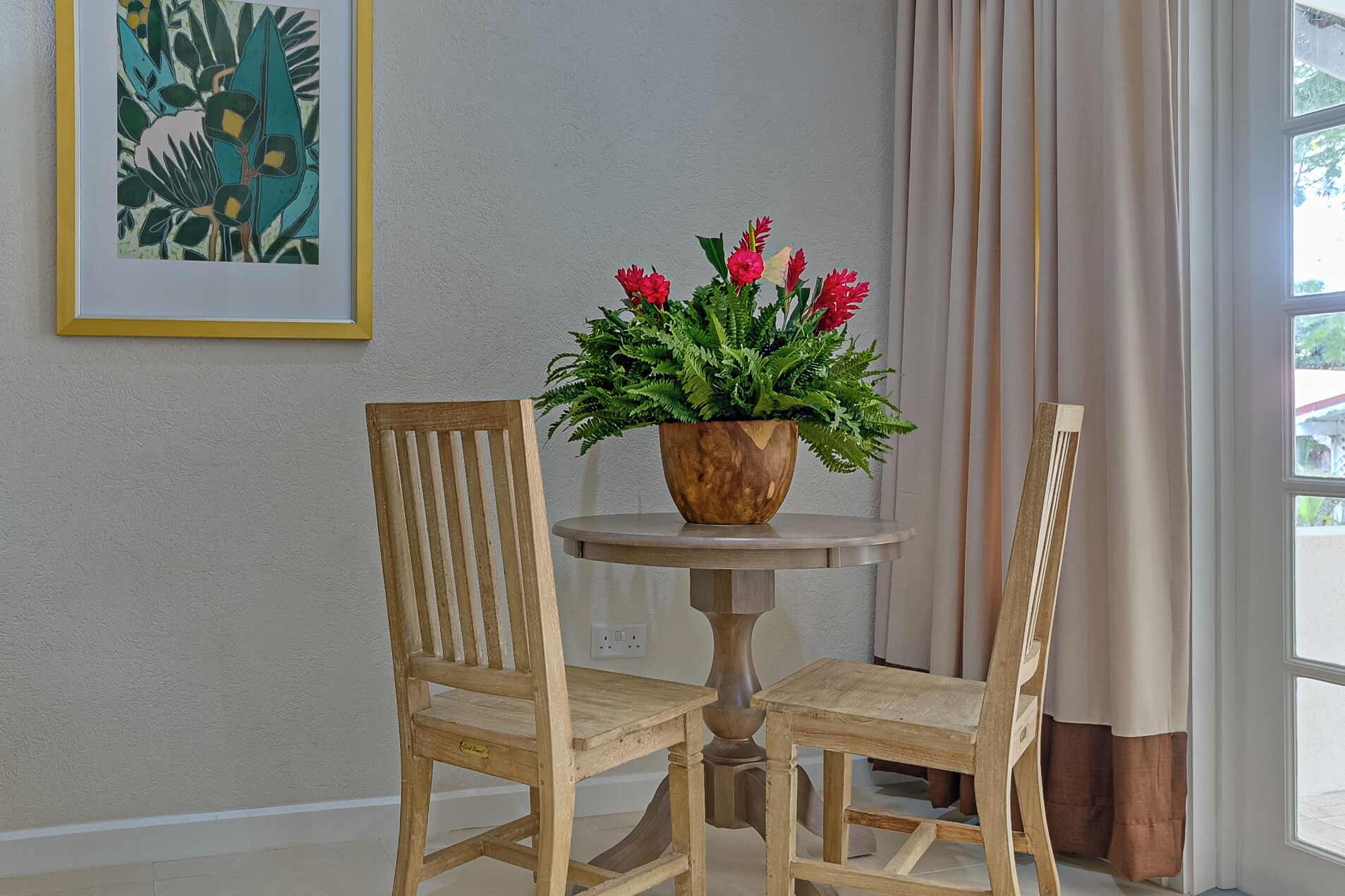 Una mesa de madera con dos sillas, un cuenco de madera con flores rojas, un cuadro en la pared y una ventana con cortinas.