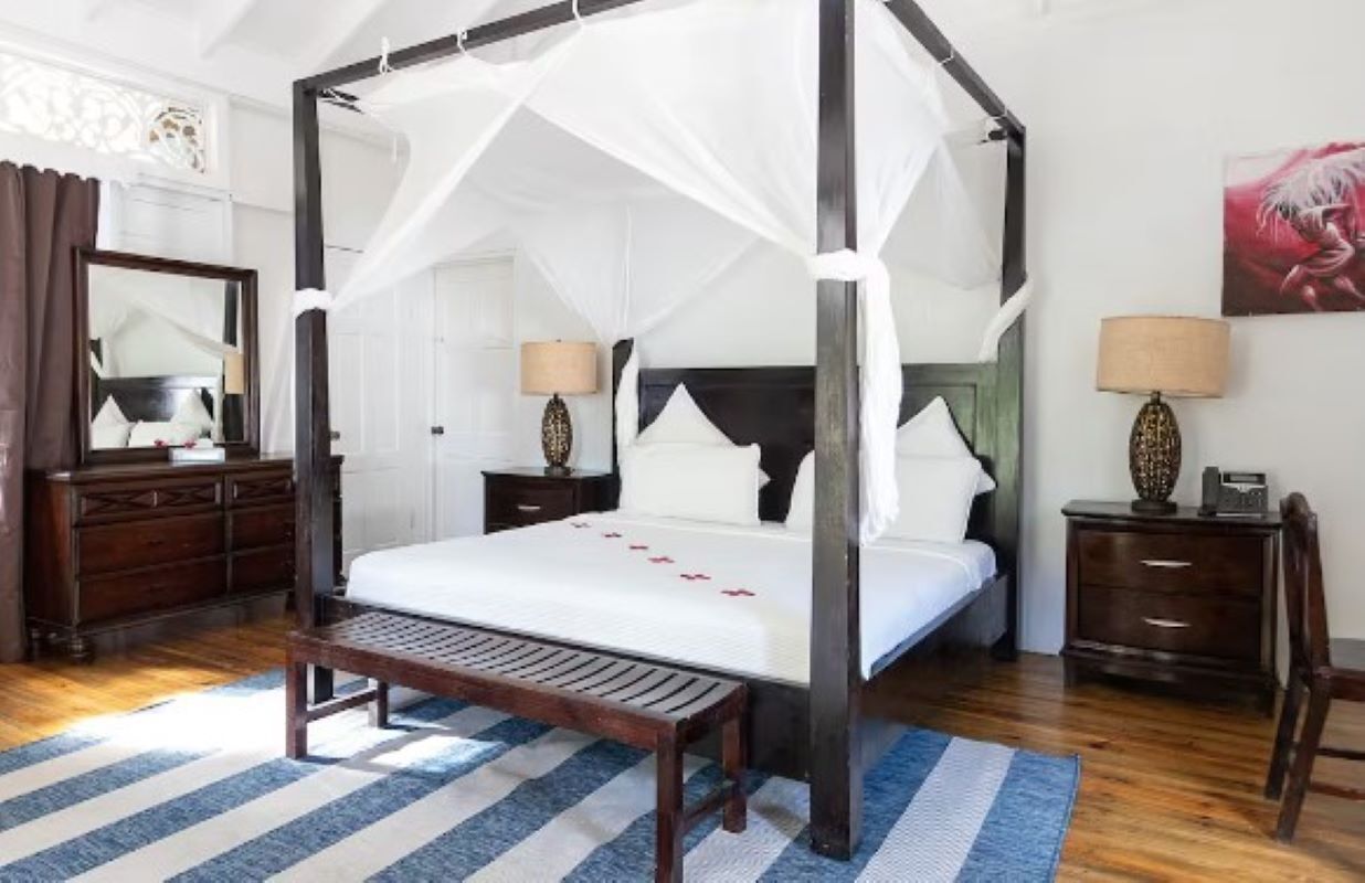 A four-poster bed with a canopy, a wooden bench, a nightstand with a lamp, and a dresser with a mirror.