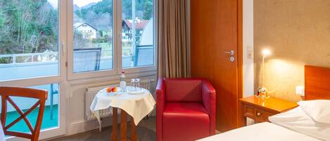 Una habitación de hotel con una cama, una silla roja, una mesita con mantel blanco y vistas al exterior a través de amplias ventanas.