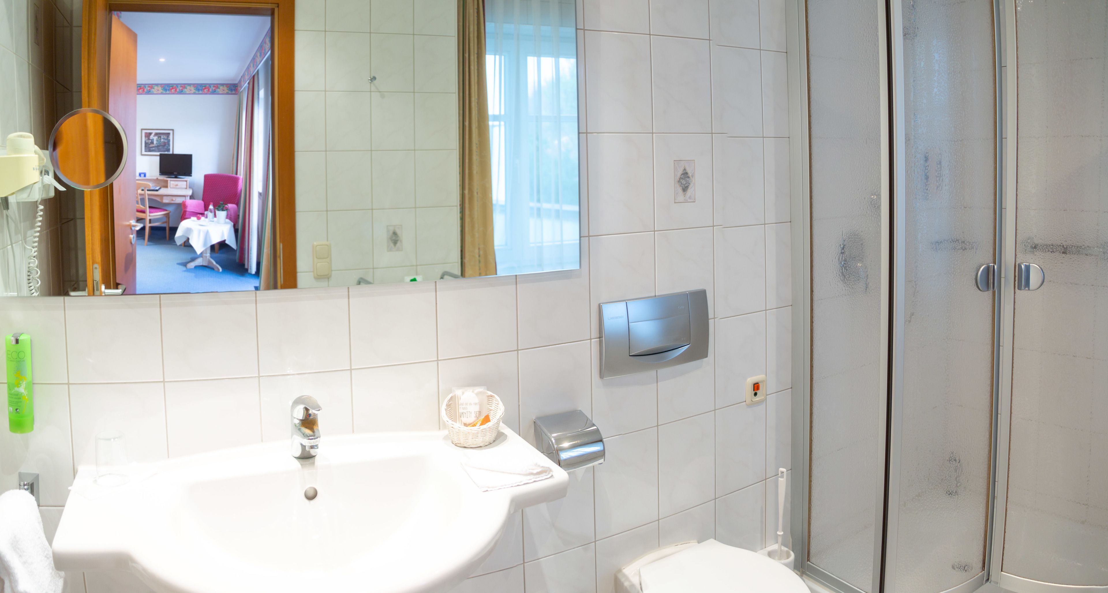 A bathroom with a sink, mirror, shower, and a view of a room with a bed and a TV.