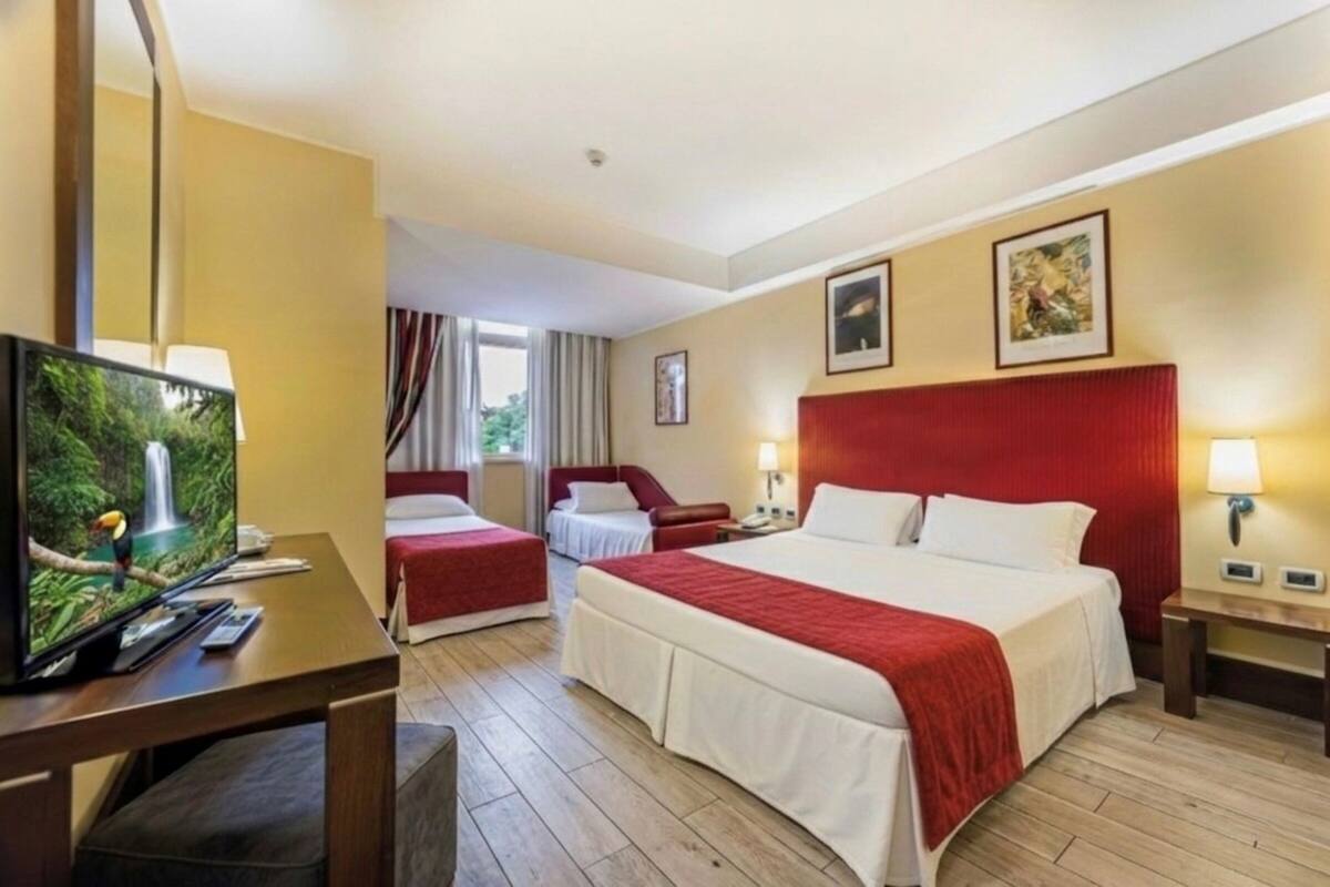 Ein Hotelzimmer mit einem großen Bett, einem kleineren Bett, einem Schreibtisch mit Fernseher und einem Fenster mit Vorhängen.