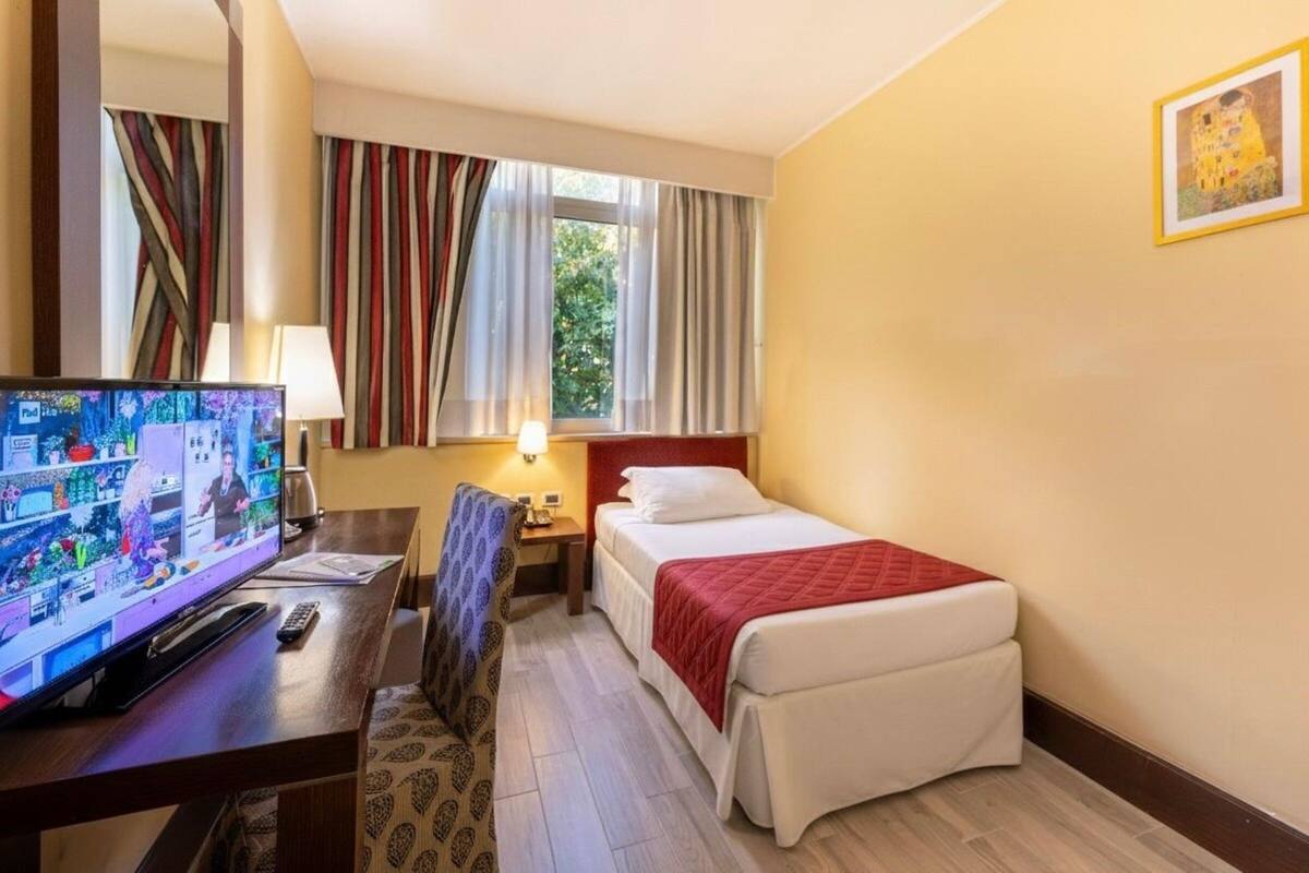 Ein Hotelzimmer mit Bett, Schreibtisch mit Fernseher, Stuhl, Nachttisch und einem fenster mit Vorhängen.