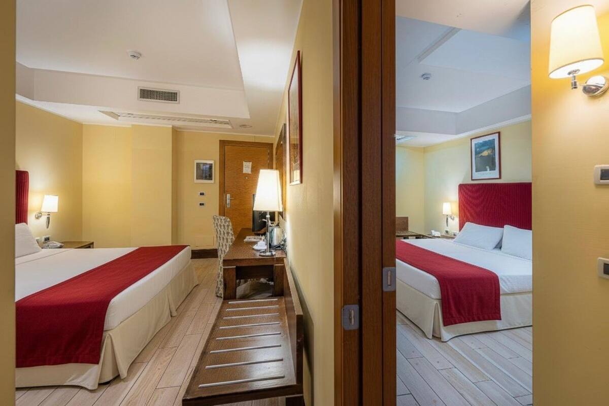 Ein Hotelzimmer mit zwei Betten, einem Schreibtisch, einem Sessel, einer Lampe und einer Tür.