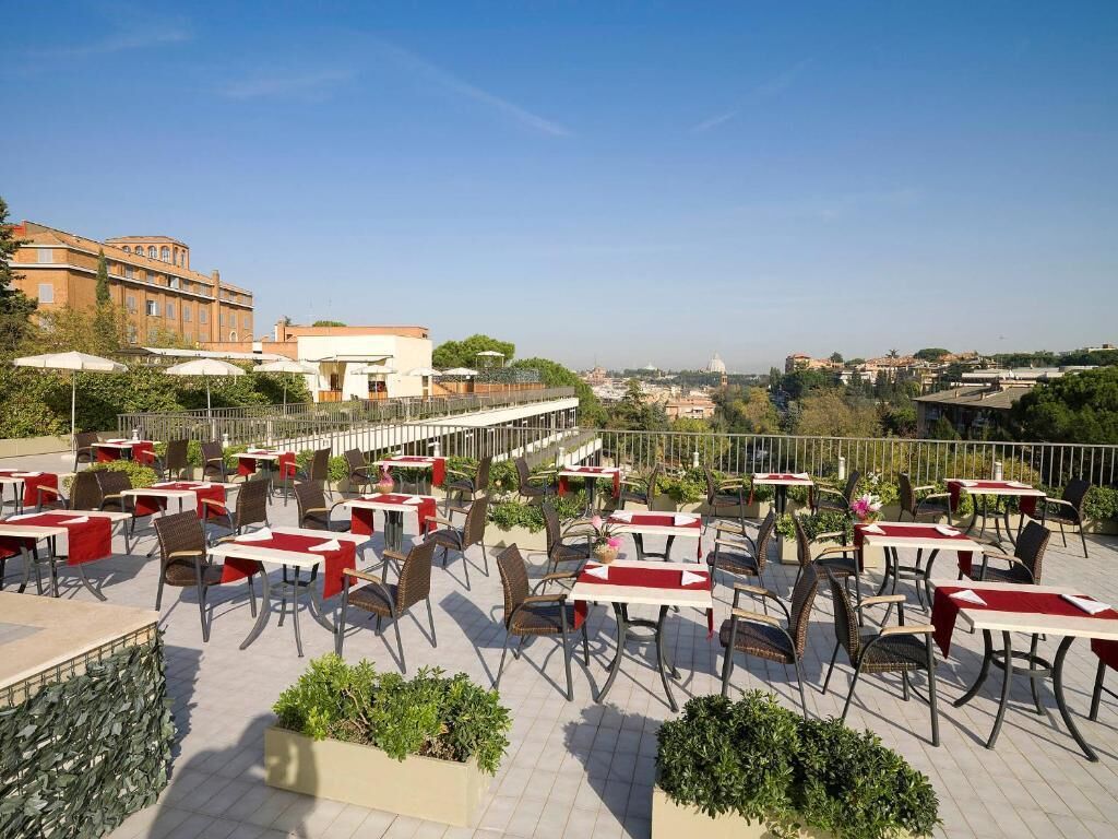 An outdoor terrace with tables and chairs, a stone wall, and a view of buildings in the distance.