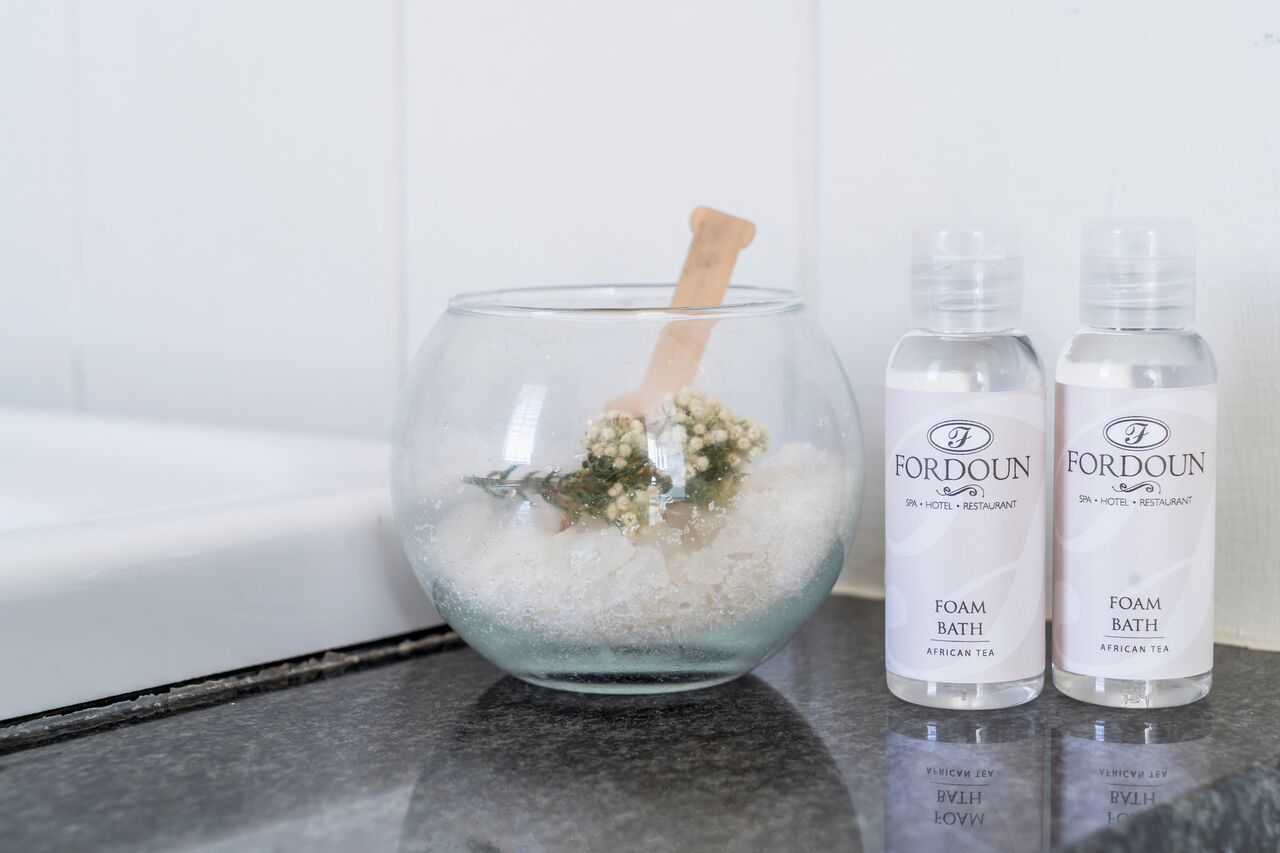 A glass bowl with a wooden spoon, two bottles of 'FORDOUN FOAM BATH AFRICAN TEA', and a bathtub in the background.