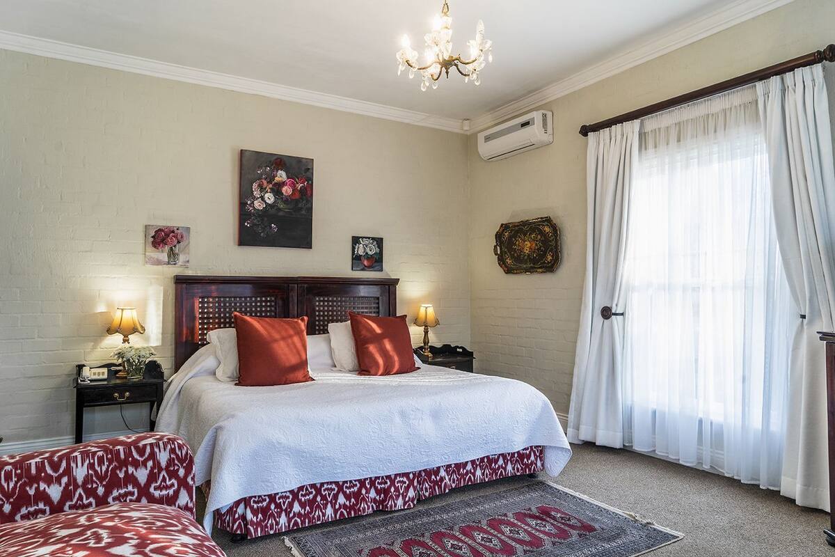 A bedroom with a bed, two bedside tables, a chandelier, an air conditioning unit, and a window with curtains.