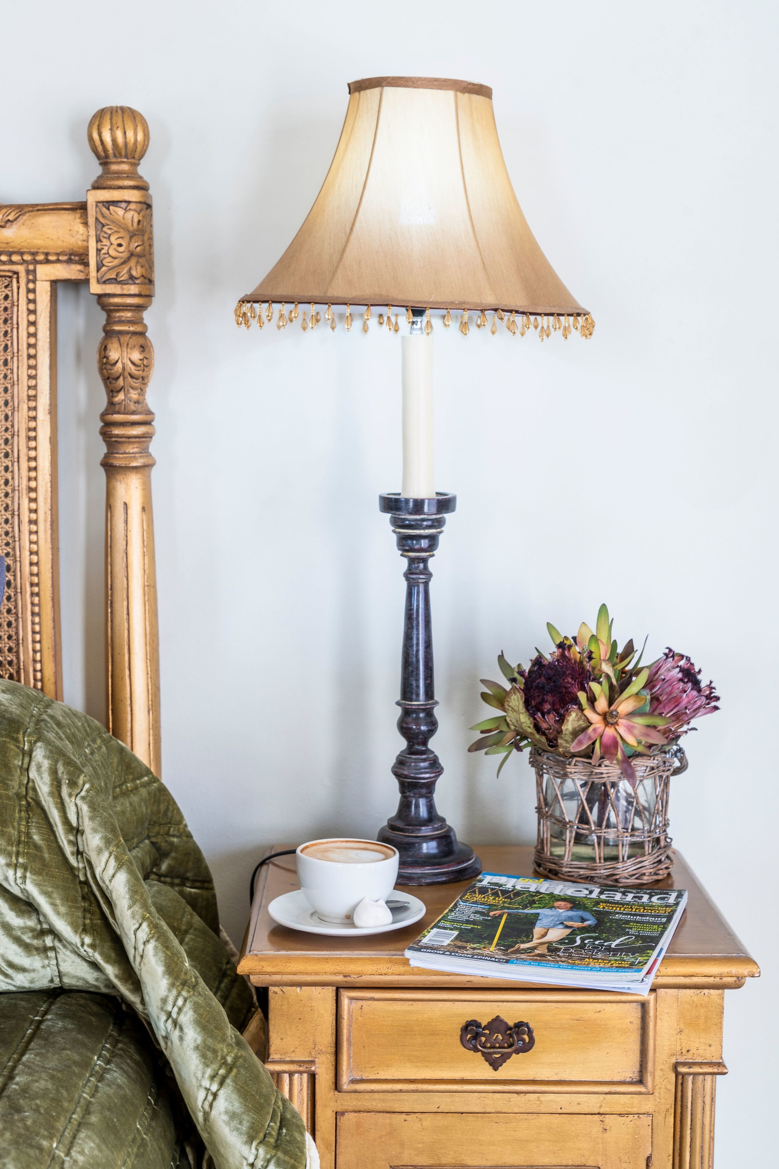 Una mesita de noche de madera con una lámpara, un florero con flores, una revista y una taza de café.