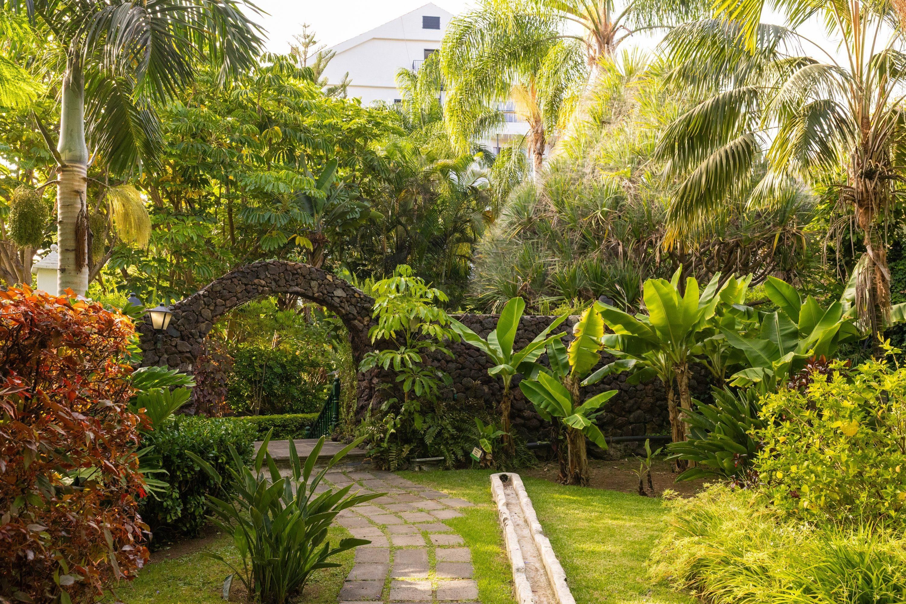Un jardín con un arco de piedra, abundante vegetación y un camino pavimentado.