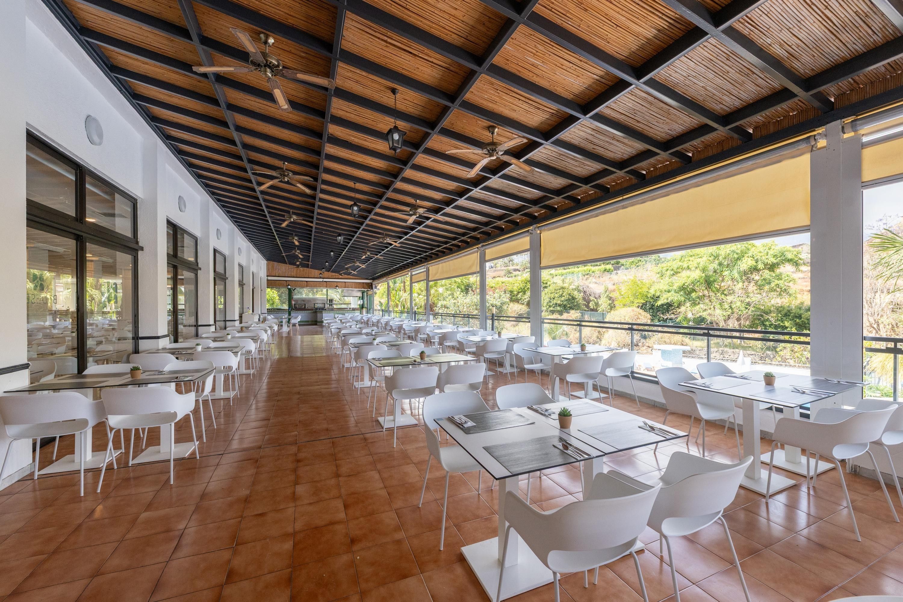 A spacious outdoor dining area with numerous tables and chairs, large windows, and a wooden ceiling structure.