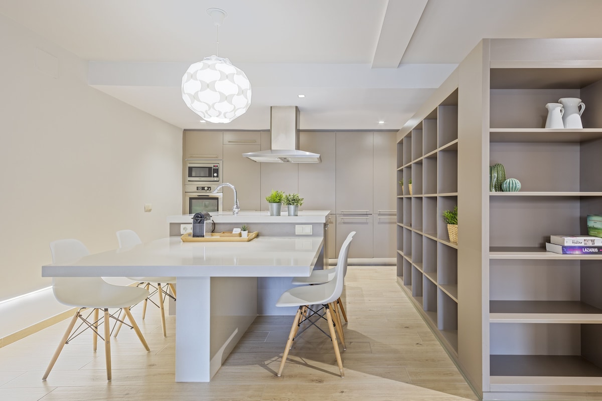 Una cocina moderna con una isla central, sillas blancas y una estantería empotrada.