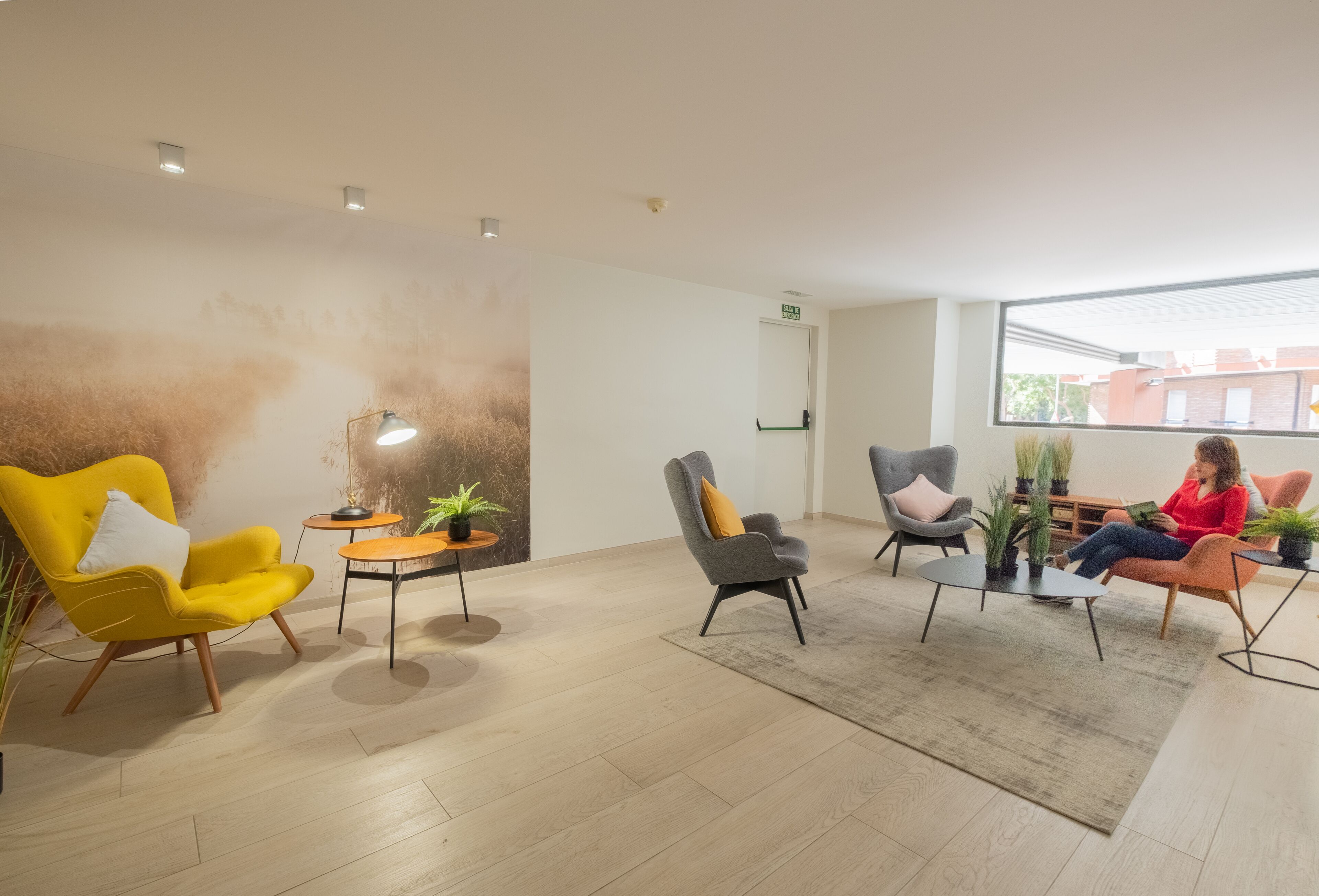 A modern waiting area with a yellow armchair, grey armchairs, and a red chair. There are potted plants and a small table with a lamp.