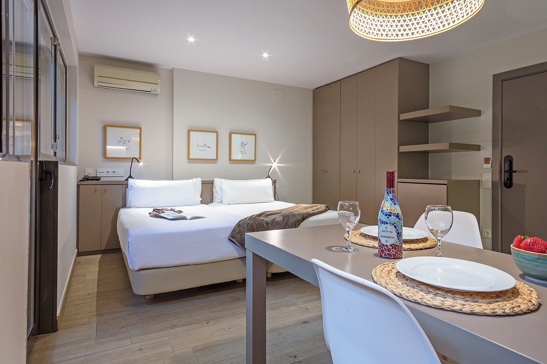 A modern hotel room with a bed, bedside table with wine glasses, a bowl of strawberries, and a minibar.