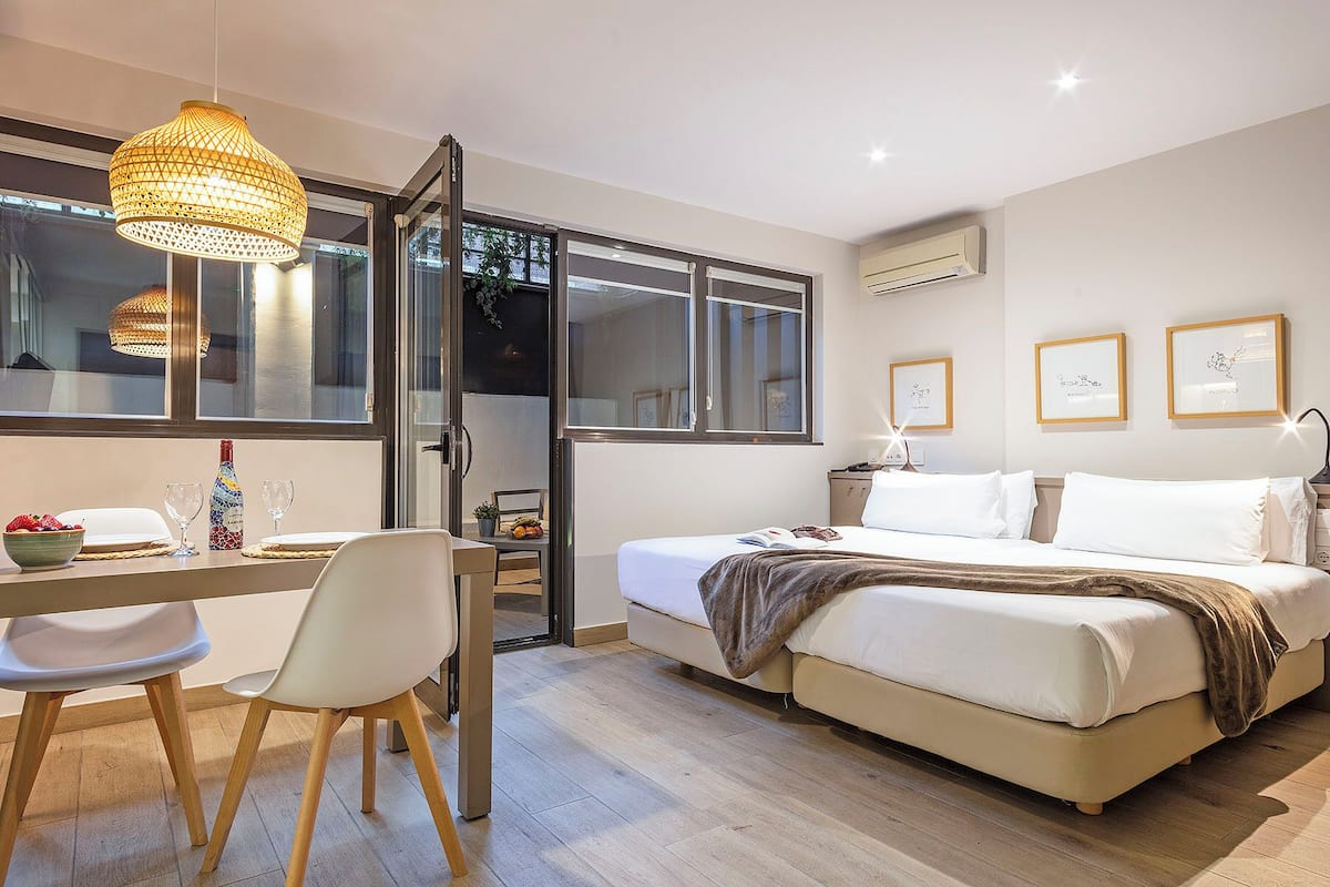 A modern hotel room with a bed, a desk with a chair, a dining table with a bowl of fruit, and a large window.
