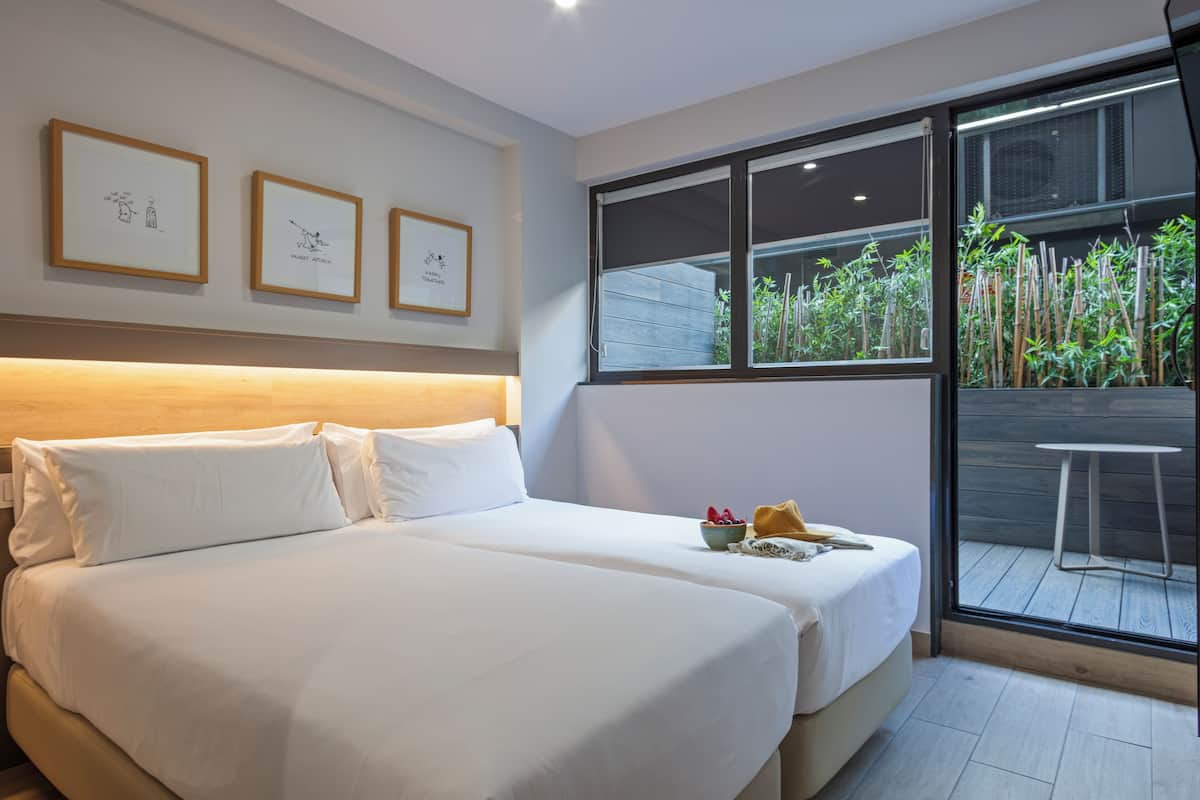 A hotel room with two beds, a large window with a view of bamboo plants, and framed artwork on the wall.