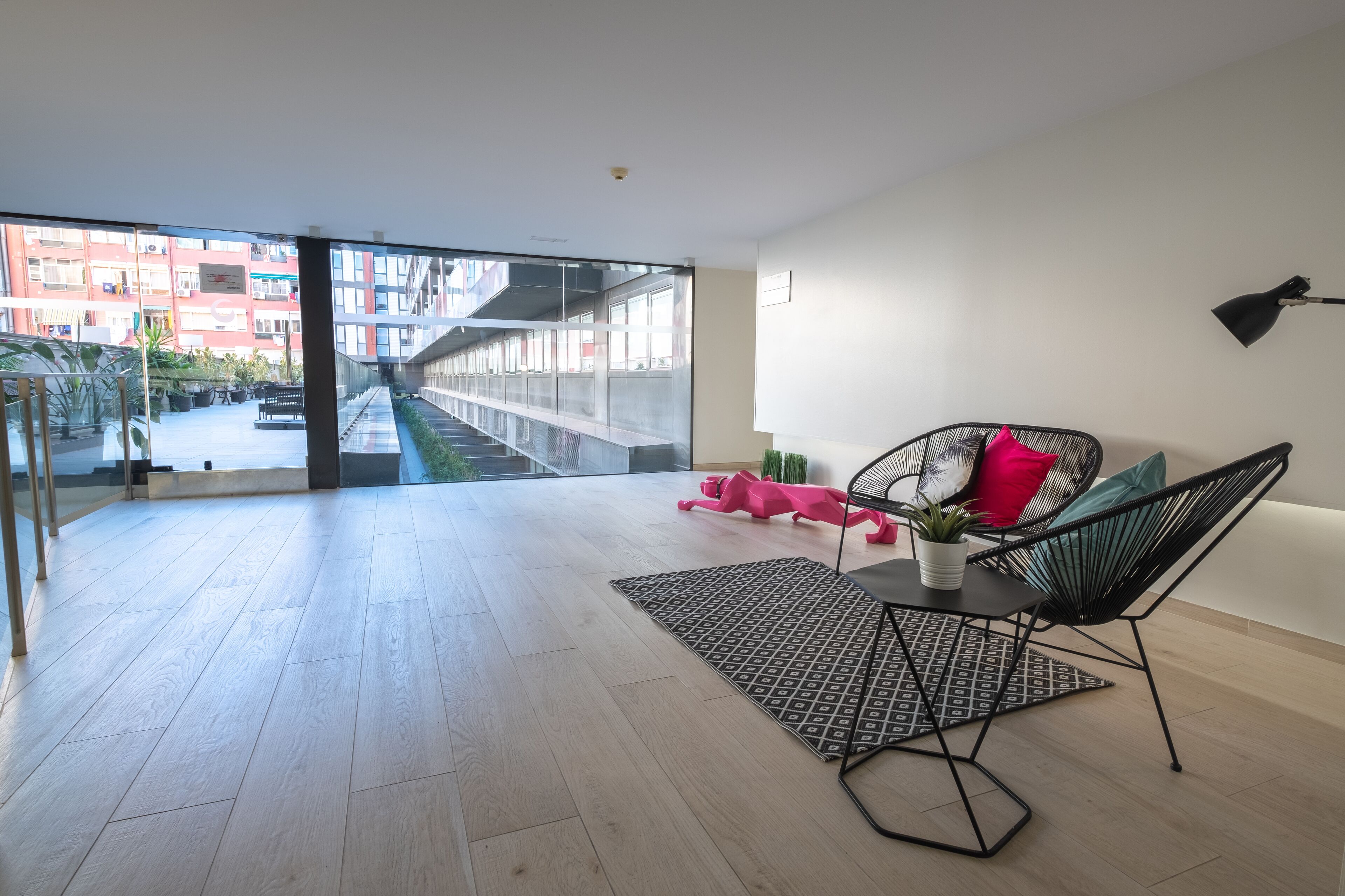 Un salon moderne doté d’une grande porte vitrée donnant sur un balcon, d’une chaise en métal noir, d’une petite table avec une plante et d’un objet décoratif rose.