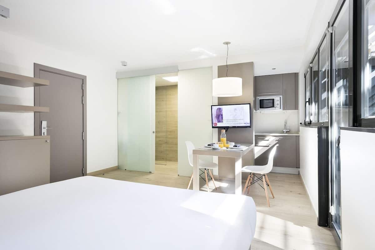 Una habitación de hotel moderna con una cama, un escritorio con computadora y una silla, un televisor y una kitchenette.