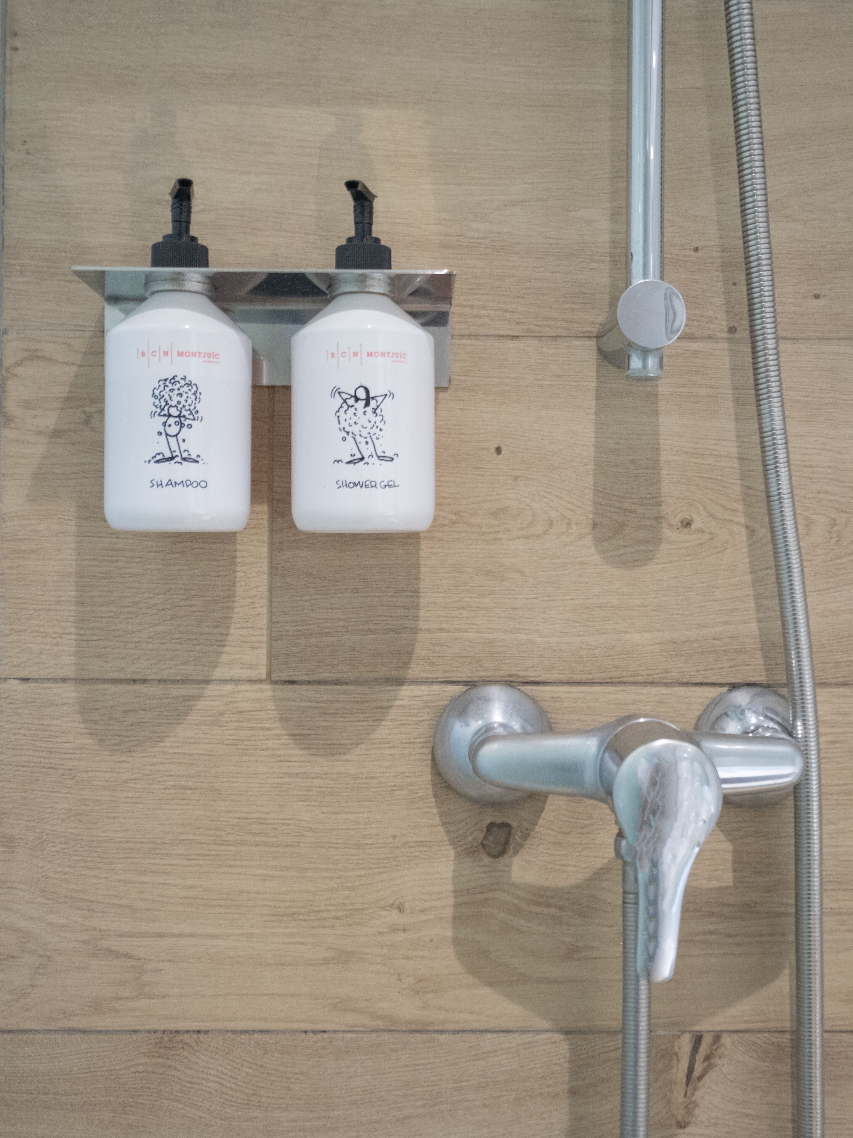 A shower area with two bottles of Montjuic shampoo and shower gel, a handheld showerhead, and a flexible hose.