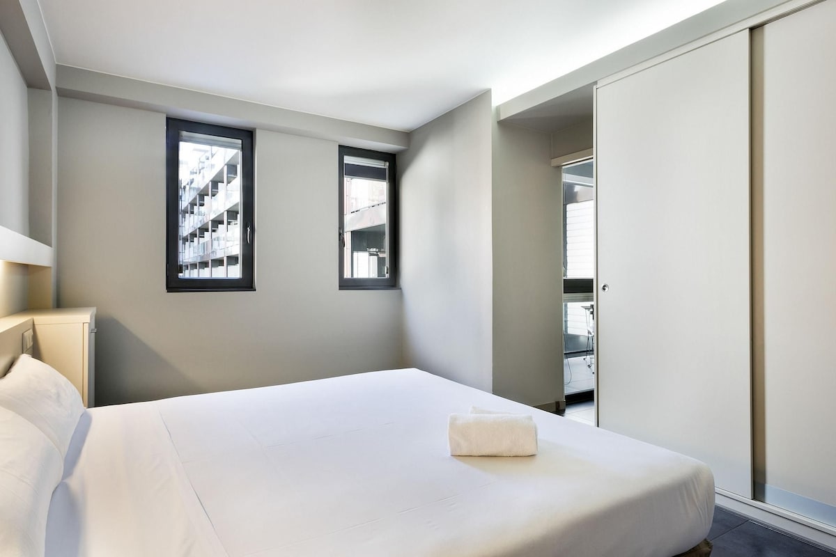 Habitación de hotel con cama, dos ventanas, armario y vista a un edificio.