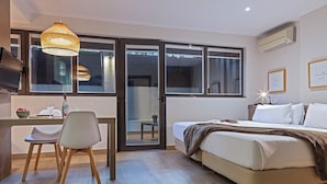 Un dormitorio moderno con una cama, un escritorio con una silla, una mesa de comedor con un cuenco de fruta y una botella, y un ventanal grande.