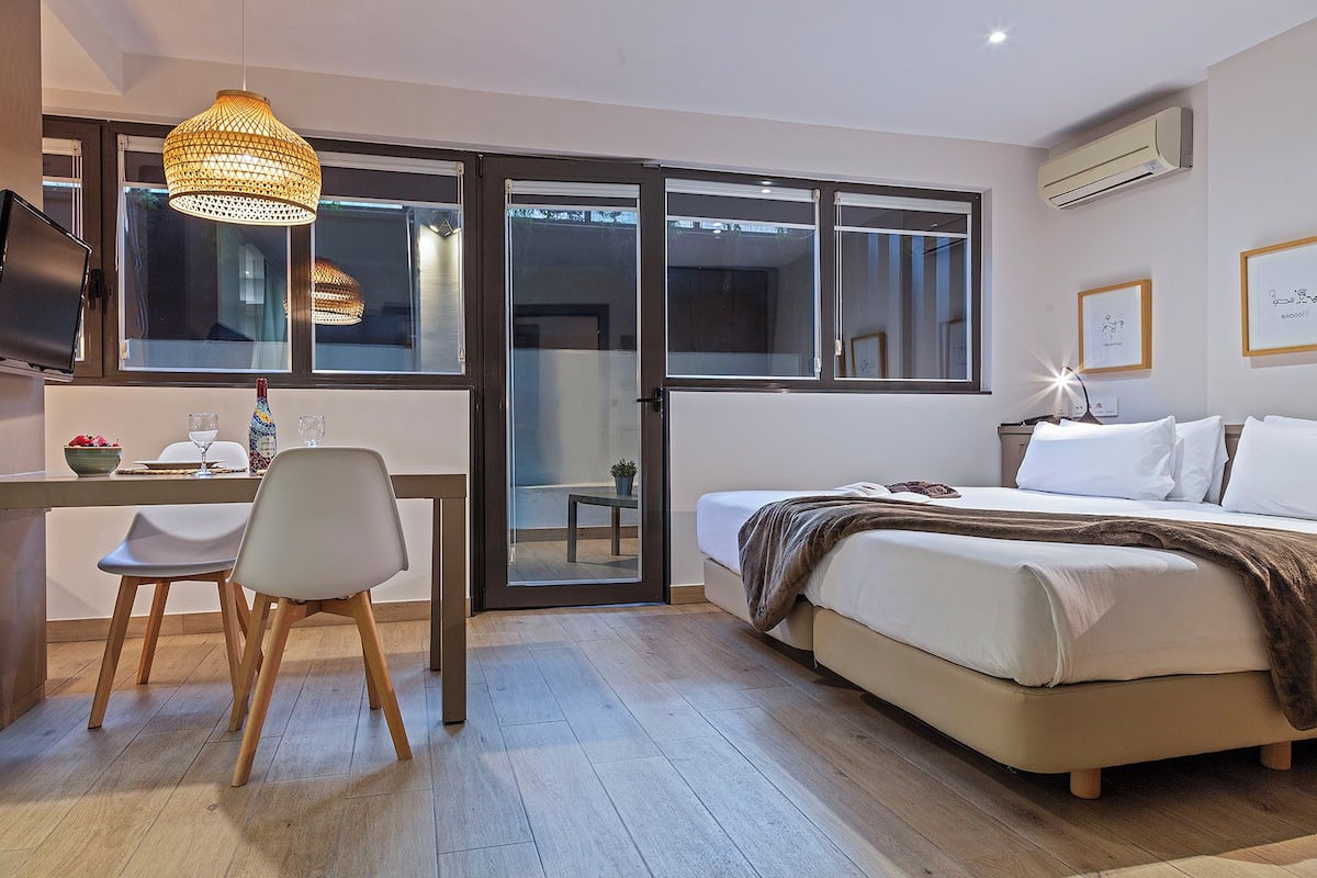 A modern bedroom with a bed, a desk with a chair, a dining table with a bowl of fruit and a bottle, and a large window.