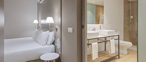 A modern hotel room with a bed, bedside lamps, a small round table, a bathroom with a shower, a sink, and a toilet.