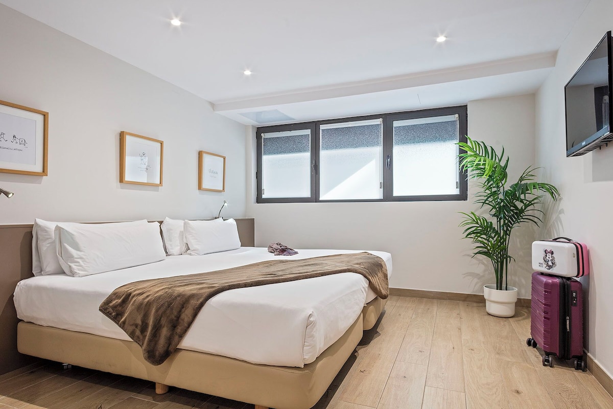 Una habitación de hotel con cama, una ventana grande, una planta en maceta y un televisor fijado a la pared.
