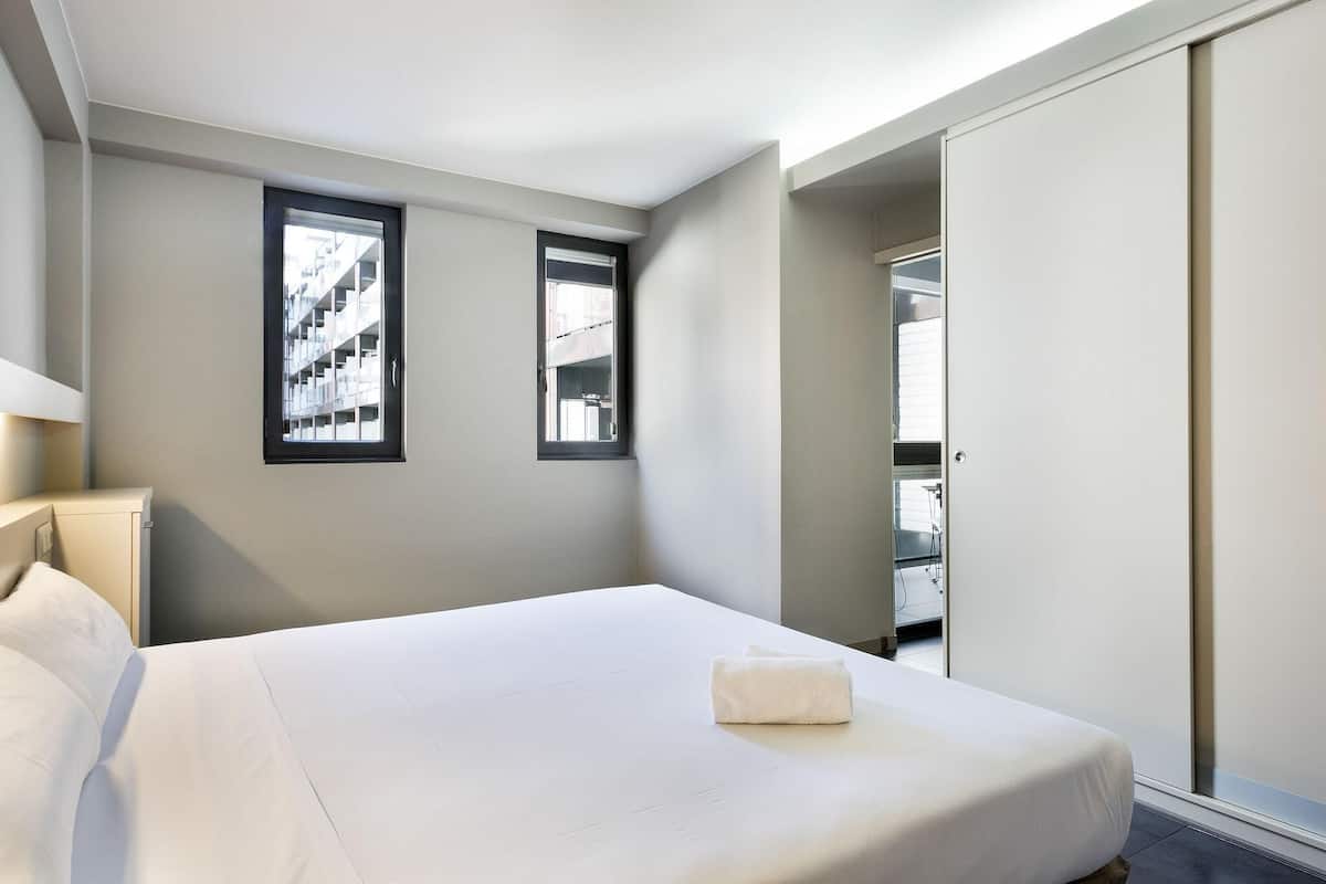 A hotel room with a bed, two windows, a wardrobe, and a view of a building outside.