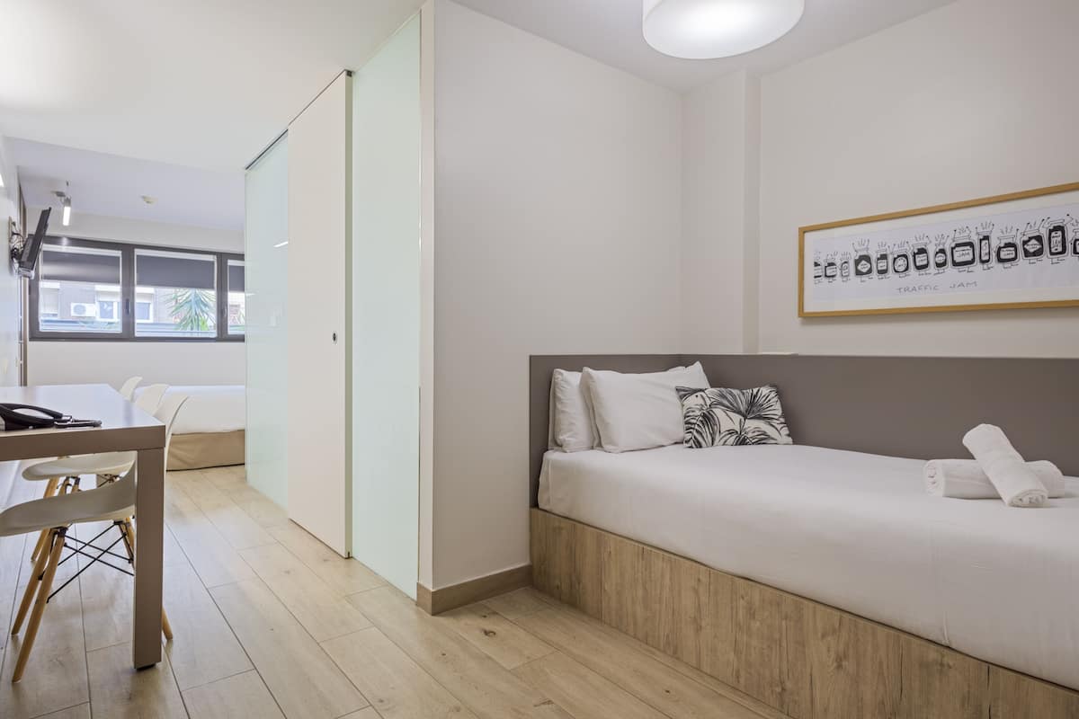 A modern hotel room with a wooden bed, white bedding, a dining table with chairs, and a view of the outside through large windows.