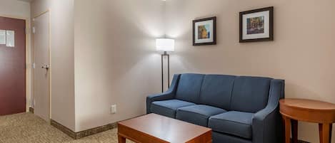 A hotel room with a blue sofa, a wooden coffee table, a floor lamp, and two framed pictures on the wall.