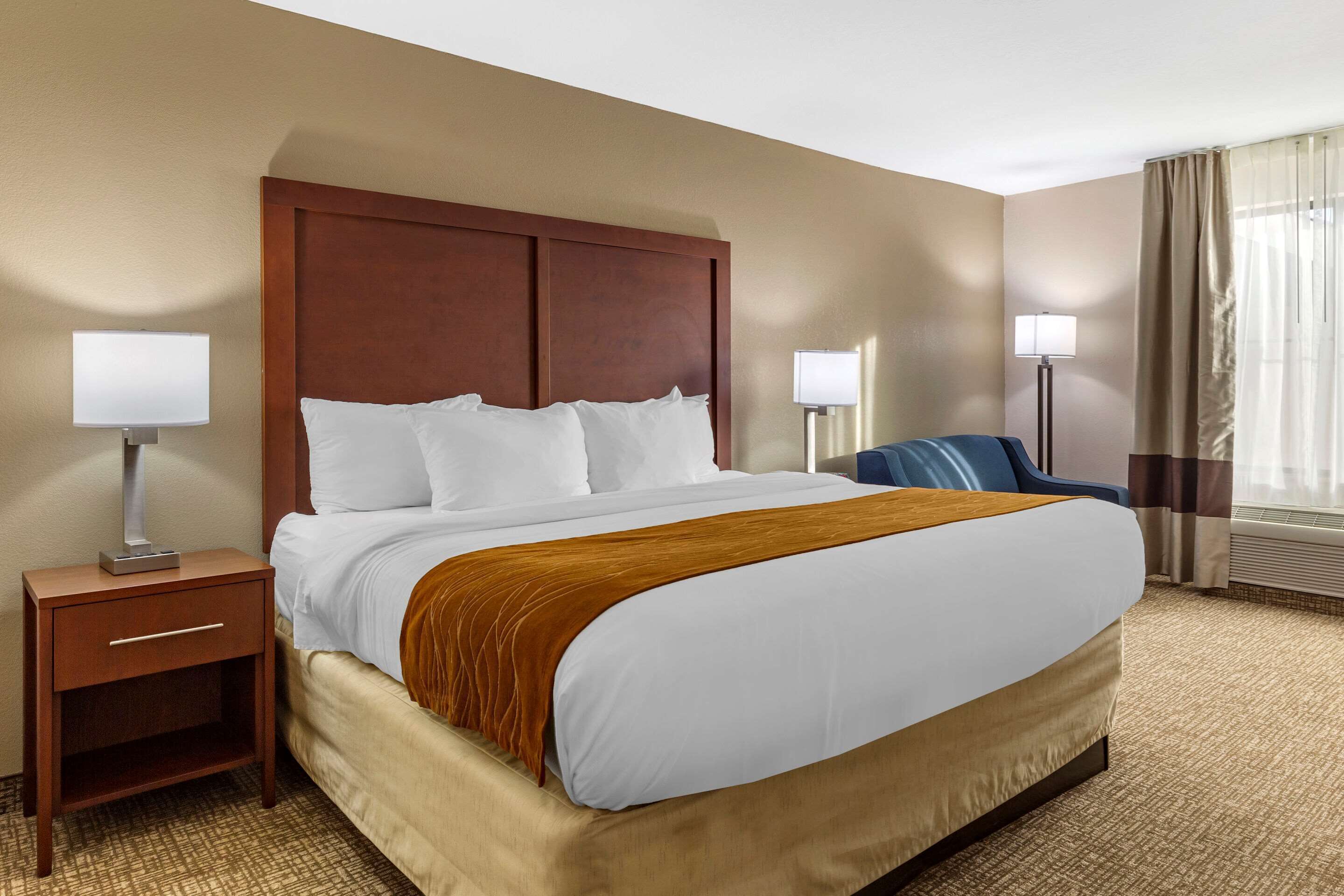 A hotel room with a large bed, a nightstand with a lamp, a chair, and a window with curtains.