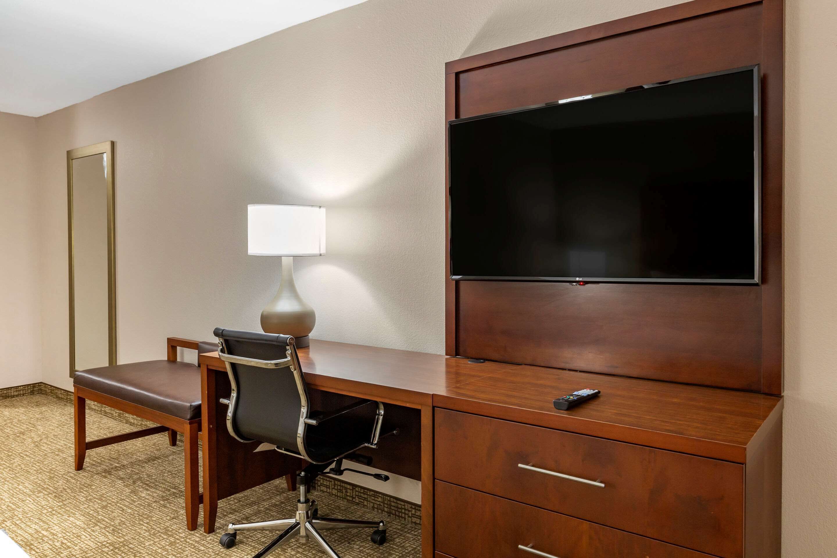 Habitación de hotel con escritorio de madera, silla de oficina negra, lámpara, televisor de pantalla plana y un espejo grande.
