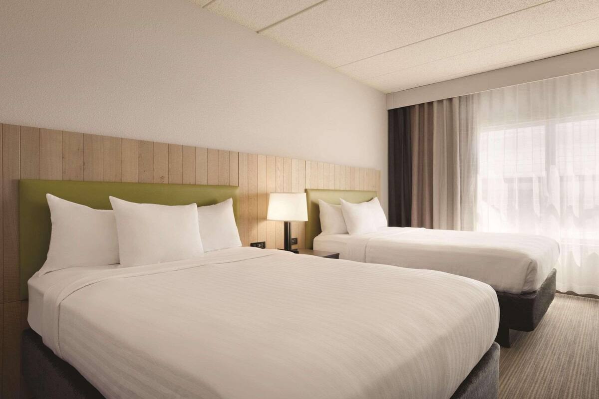 A hotel room with two beds, a wooden headboard, a nightstand with a lamp, and a large window with curtains.