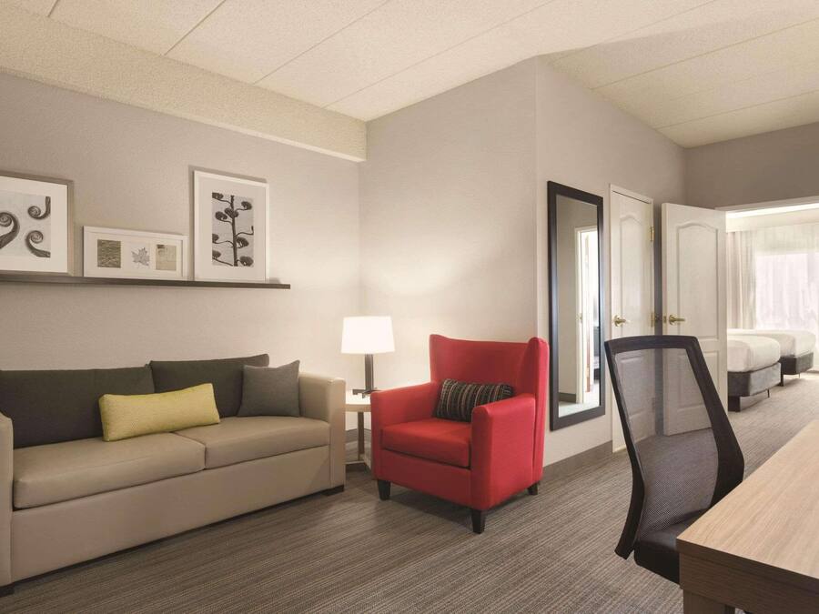 A hotel room with a sofa, a red armchair, a desk, and a bed. There are framed pictures on the wall and a window with curtains.